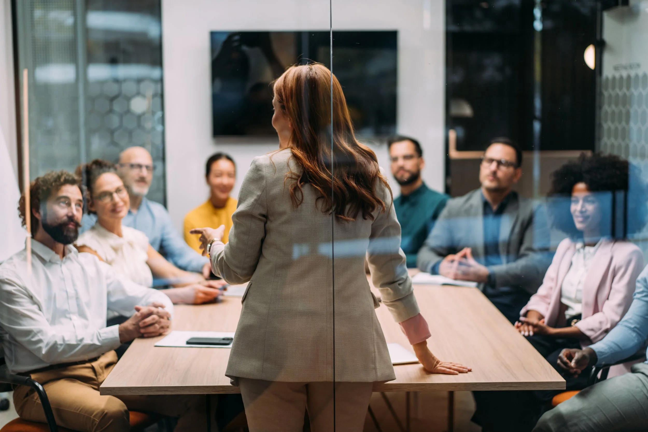 A woman with long brown hair, dressed in a gray blazer, is giving a presentation to a diverse group of professionals seated around a conference table in a modern office conference room. The group is attentively listening.
