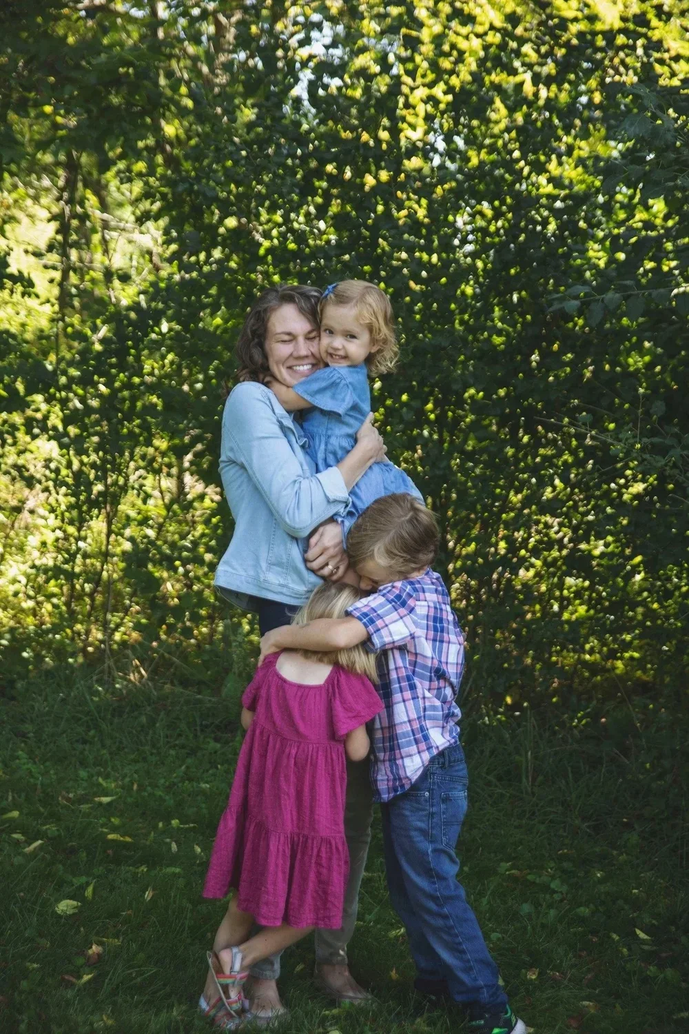 A woman with three children hugging outdoors near green trees, smiling and happy.