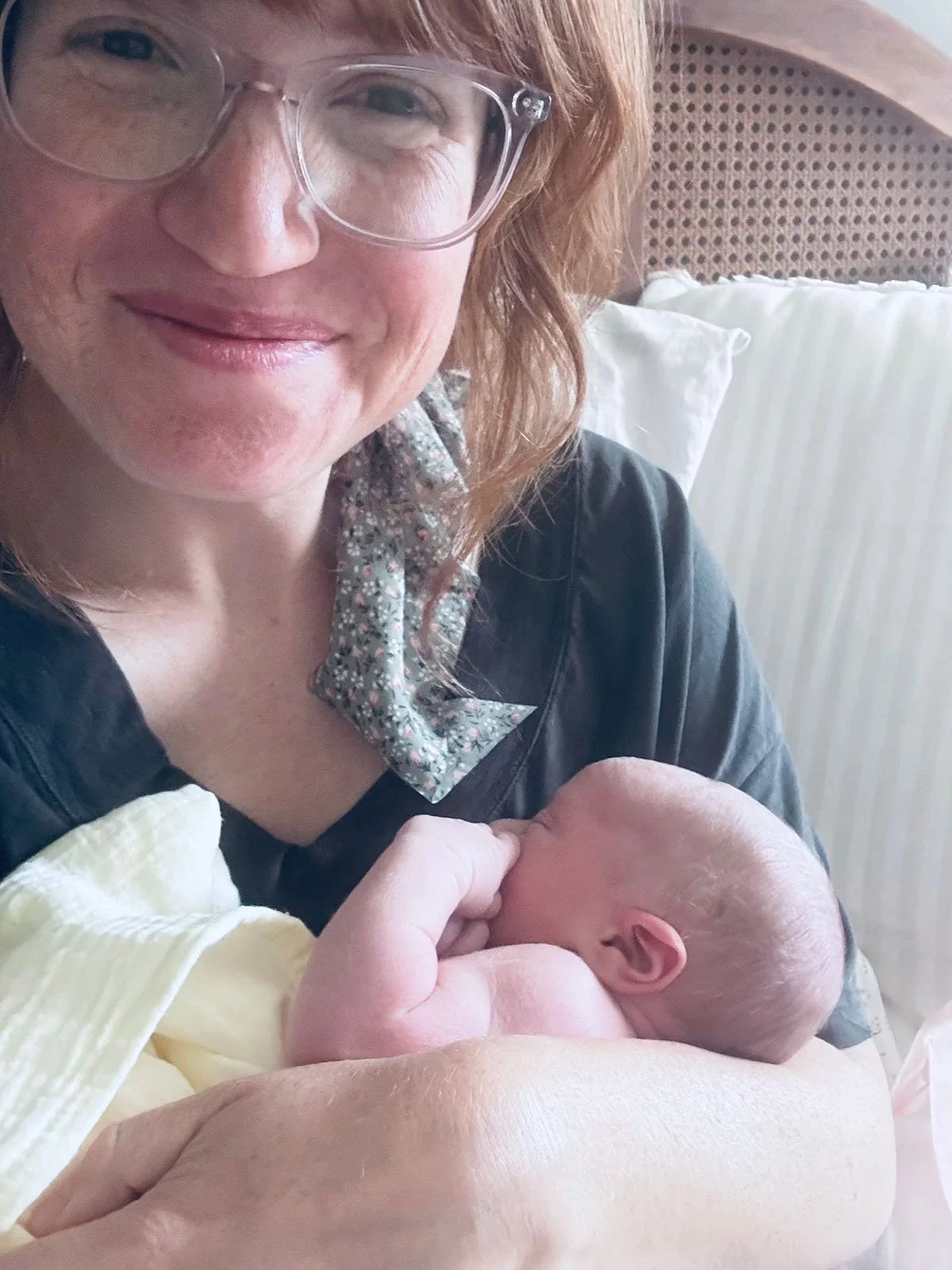 A woman with glasses and curly red hair holding a newborn baby in a hospital room.
