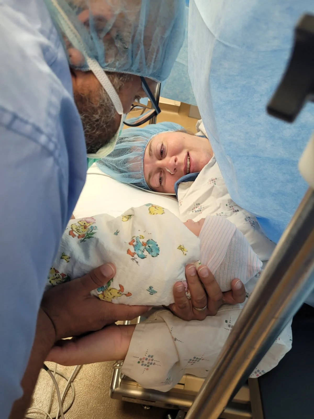 A couple of medical professionals, dressed in protective gear, holding a newborn baby in a hospital room. The mother is lying in a hospital bed and smiling at her baby, while the professionals carefully cradle the infant.