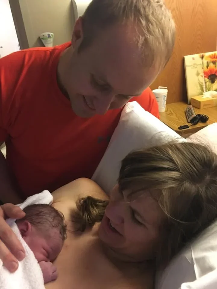 A woman lying in bed holding a newborn baby, with a man leaning over her, all smiling in a warmly lit room.