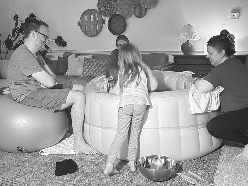 Family gathering in a living room, with children and adults sitting around a round, padded playpen, some engaging with children, with decorative wall art and a table lamp in the background.
