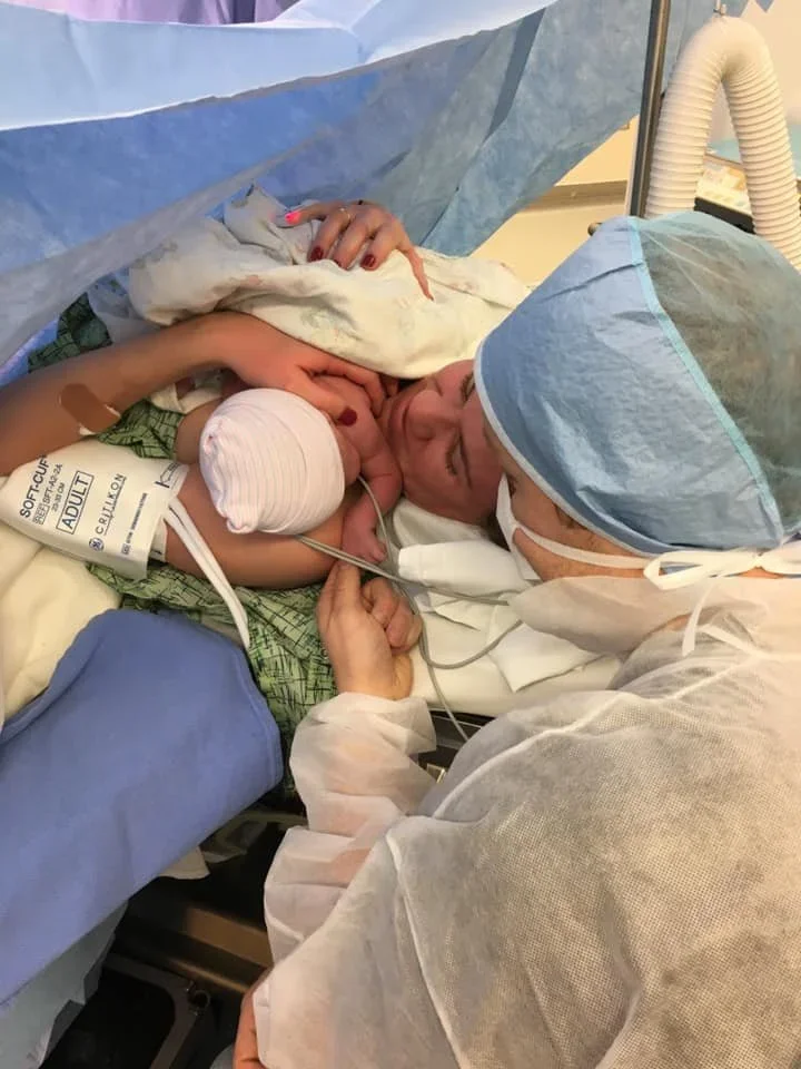 A woman holding a newborn baby in a hospital delivery room, connected to medical equipment with a healthcare worker nearby.