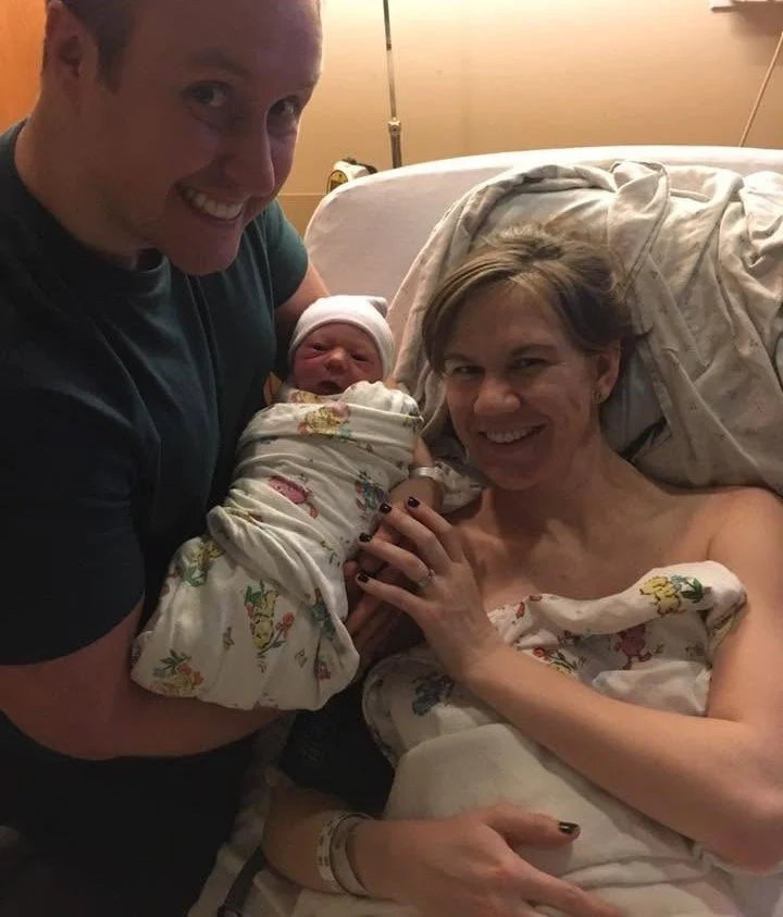 A man is holding a newborn baby wrapped in a hospital blanket next to a woman lying in a hospital bed, also wrapped in a hospital blanket, smiling at the camera.