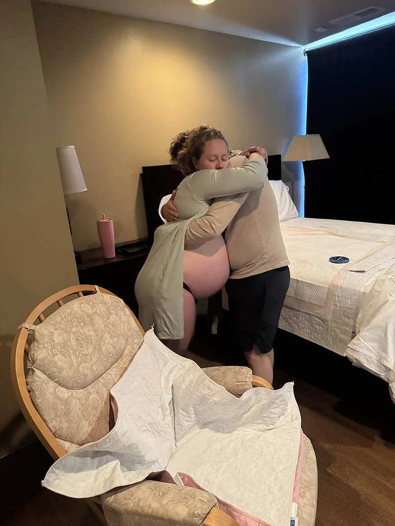 A woman and a man hugging each other in a hotel room. The woman is partially covered with a blanket and the man is wearing a beige shirt and black shorts. There is an empty beige armchair with a white cloth draped over it in the foreground, and a bed with a beige cover in the background. The room has a beige wall, a bedside table with a lamp, and dark curtains with blue lighting.