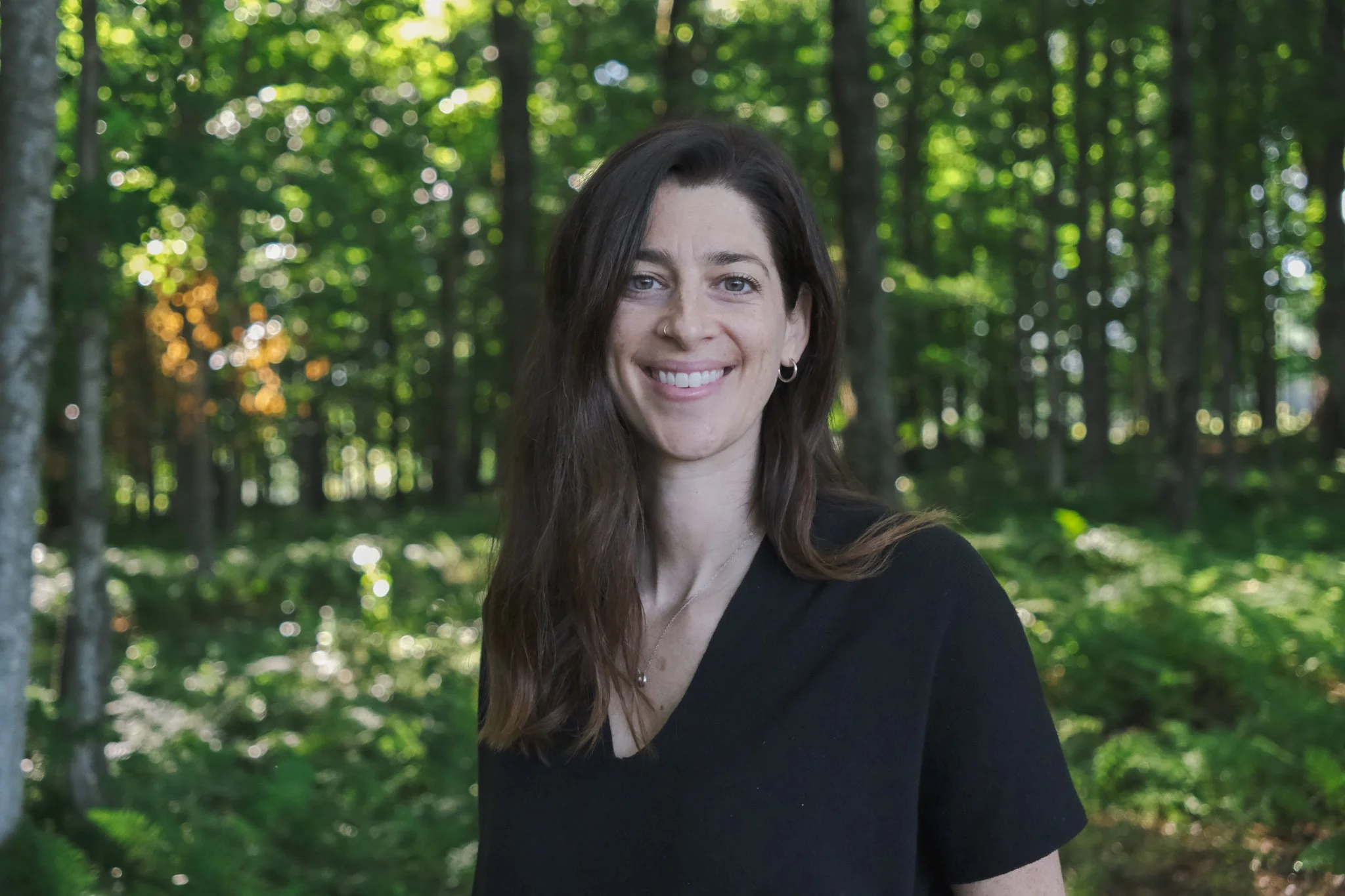 Portrait d'une jeune femme avec des cheveux bruns, portant un collier, souriant, dans une forêt verdoyante.