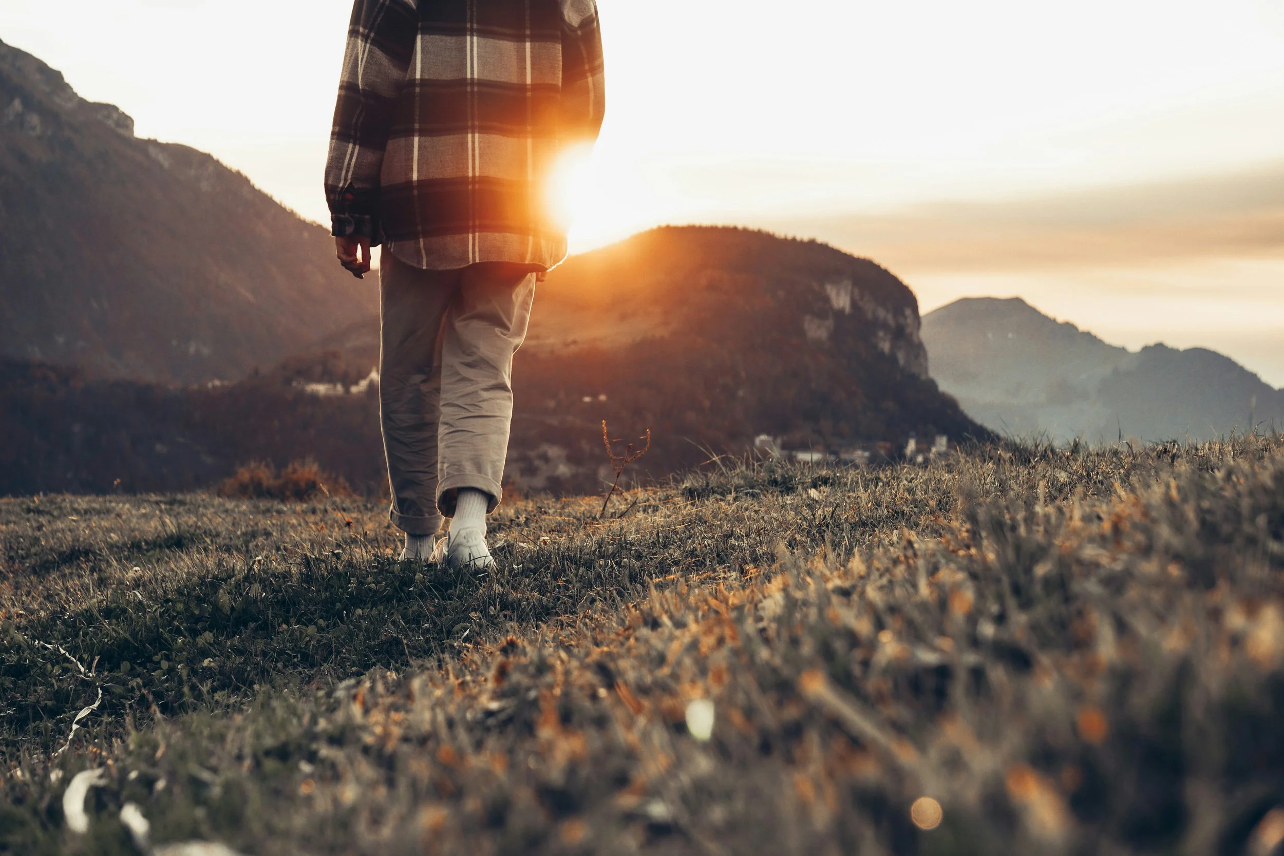 Une personne marche dans un champ avec des montagnes en arrière-plan, au coucher du soleil, portant un pull à carreaux et un pantalon beige.