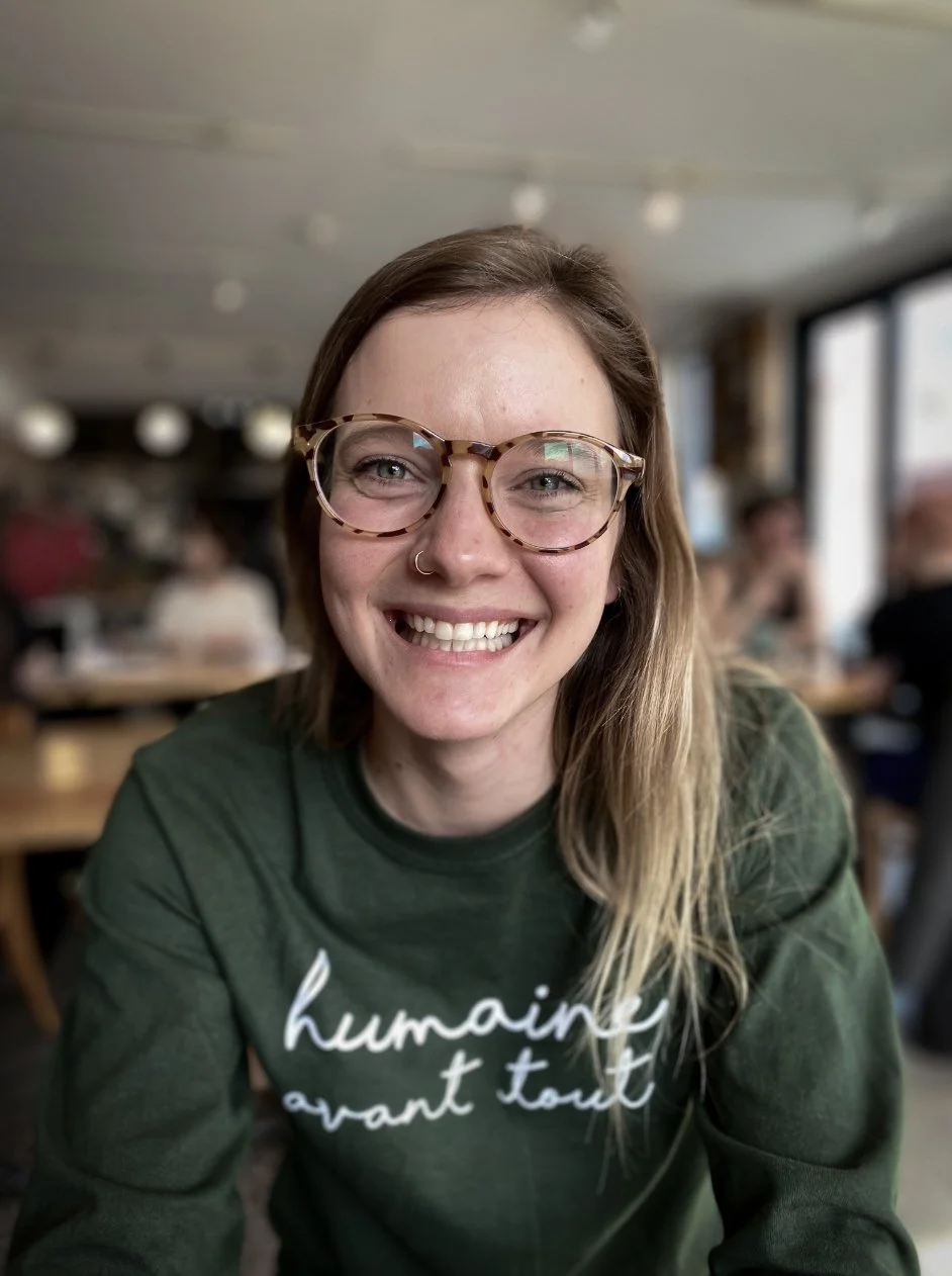 Jeune femme souriante portant des lunettes à la mode, avec un t-shirt vert, assise à une table dans un café, ambiance chaleureuse.