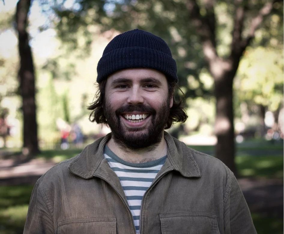 Un homme souriant avec une barbe portant un bonnet noir, une veste beige et un t-shirt à rayures, dans un parc avec des arbres en arrière-plan.