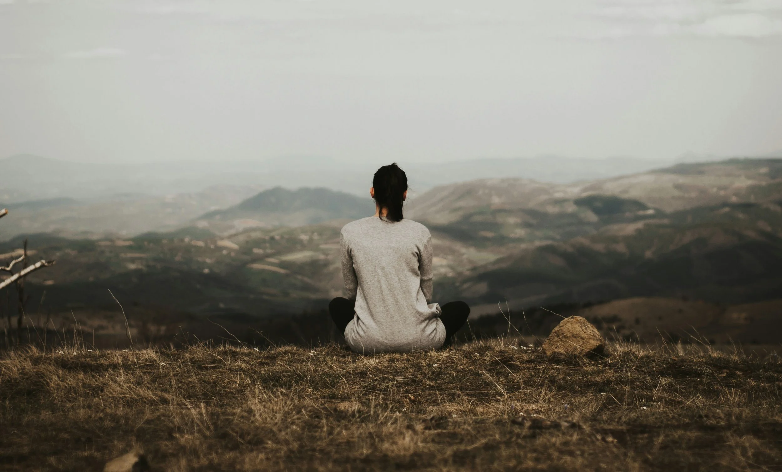 Une personne assise en position de lotus, regardant un paysage de montagnes et de vallées sous un ciel nuageux.