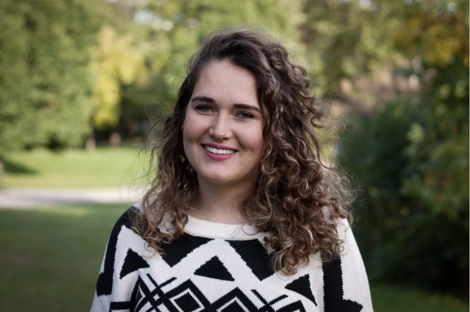 Jeune femme souriante avec des cheveux bouclés, portant un pull noir et blanc, dans un parc en journée.