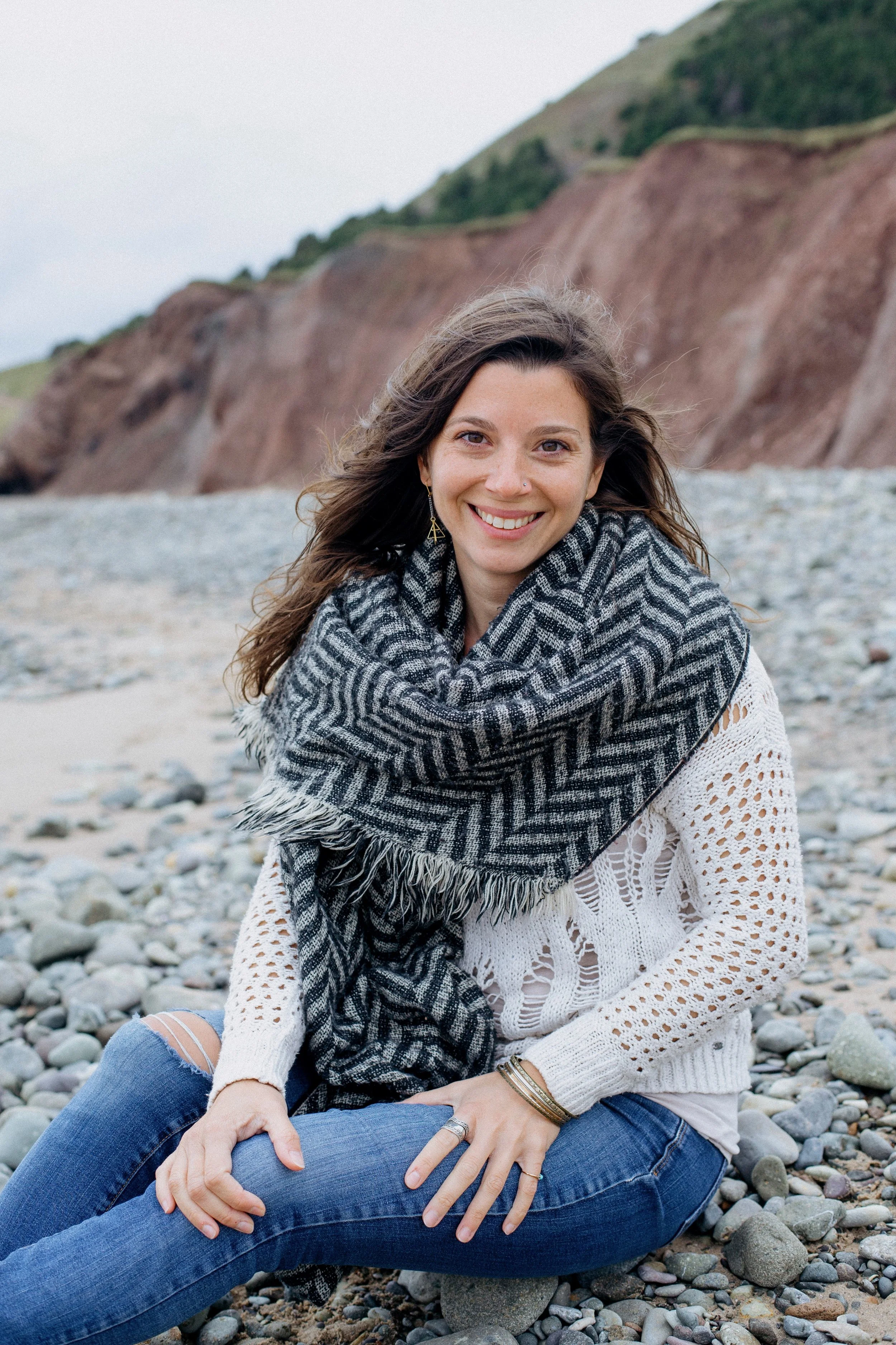 Jeune femme souriante assise sur une plage de galets, vêtue d'une chemise en crochet blanche, un foulard gris à motifs, et portant des bracelets, avec des falaises en arrière-plan.
