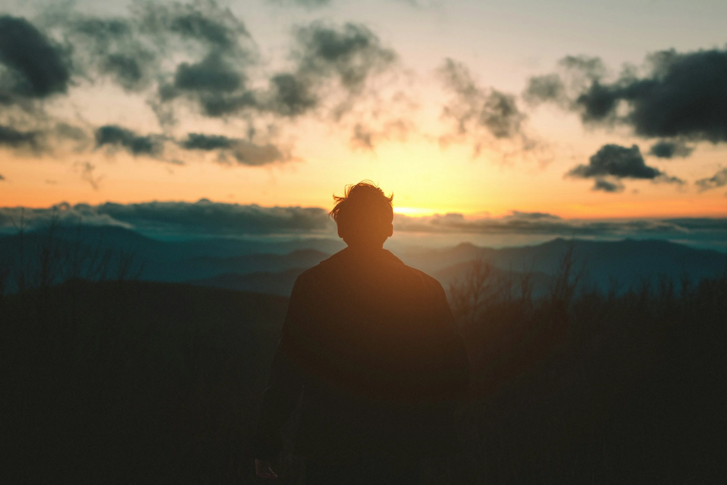 Un silhouette d'une personne regardant un coucher de soleil à l'horizon, avec un ciel rempli de nuages.