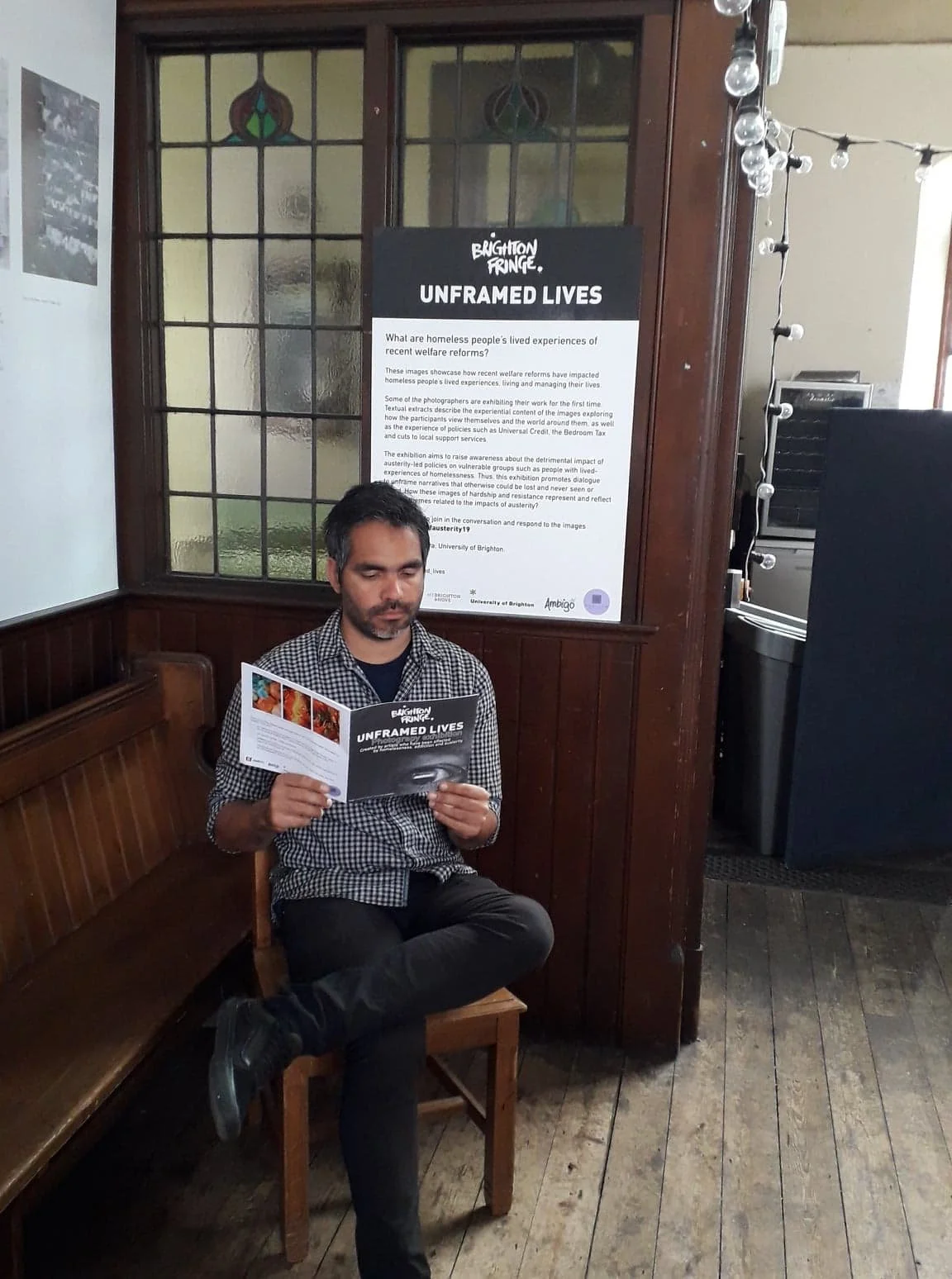 A man sitting on a wooden bench in a room with wood-paneled walls, reading a brochure titled "Brighton Fringe Unframed Lives," with a large informational poster about the event on the wall behind him. The room has stained glass windows and string lights.