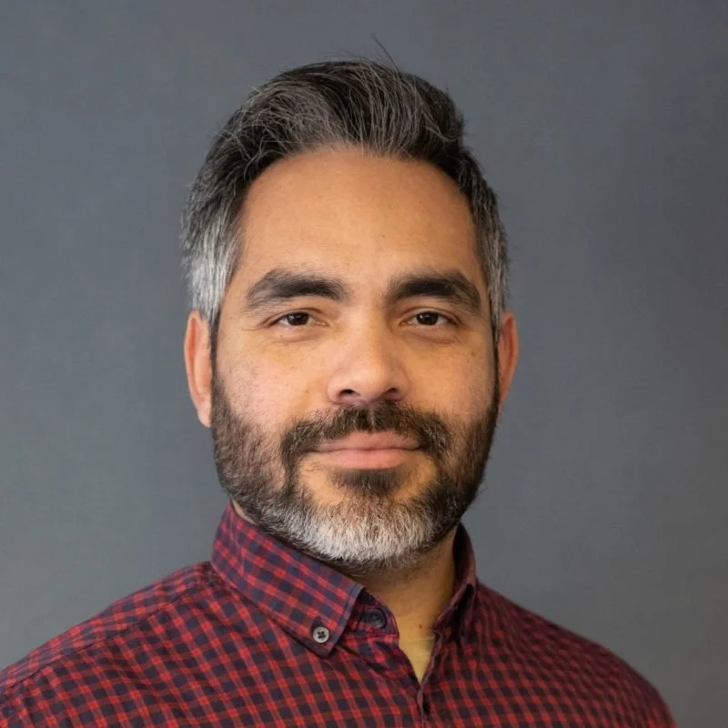 Headshot of a man with salt-and-pepper hair and beard, wearing a red and black checkered shirt against a gray background.