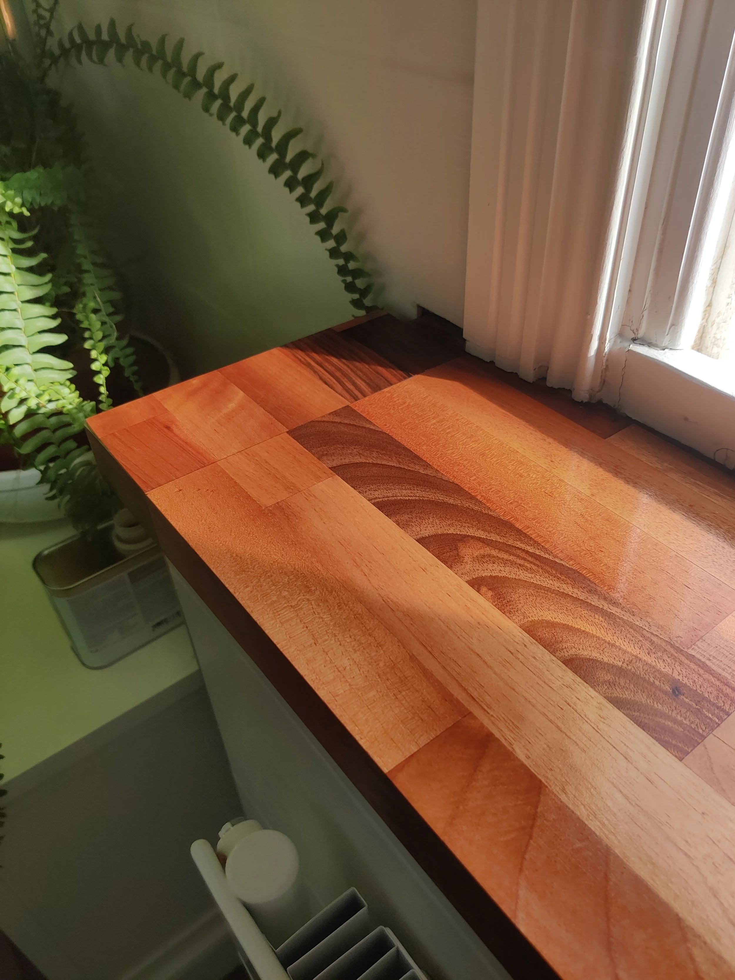 A corner of a wooden window board with reddish-brown oak wood grain, next to a white wall with a window and white curtains. To the left, there are green leafy plants and a small metal container.