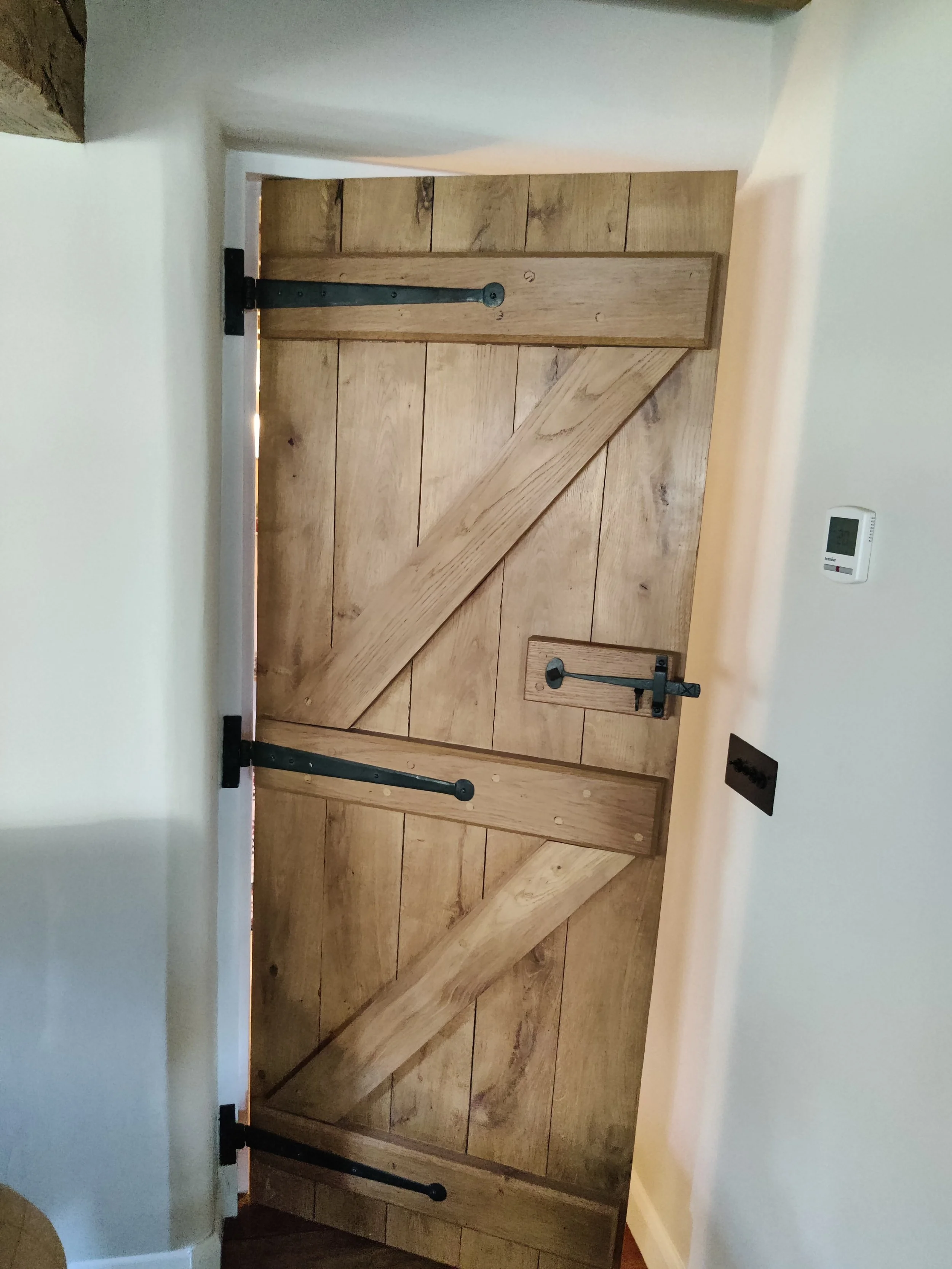 A rustic wooden oak door with black metal hinges and latch, set against a white wall with a thermostat and electrical outlet nearby.