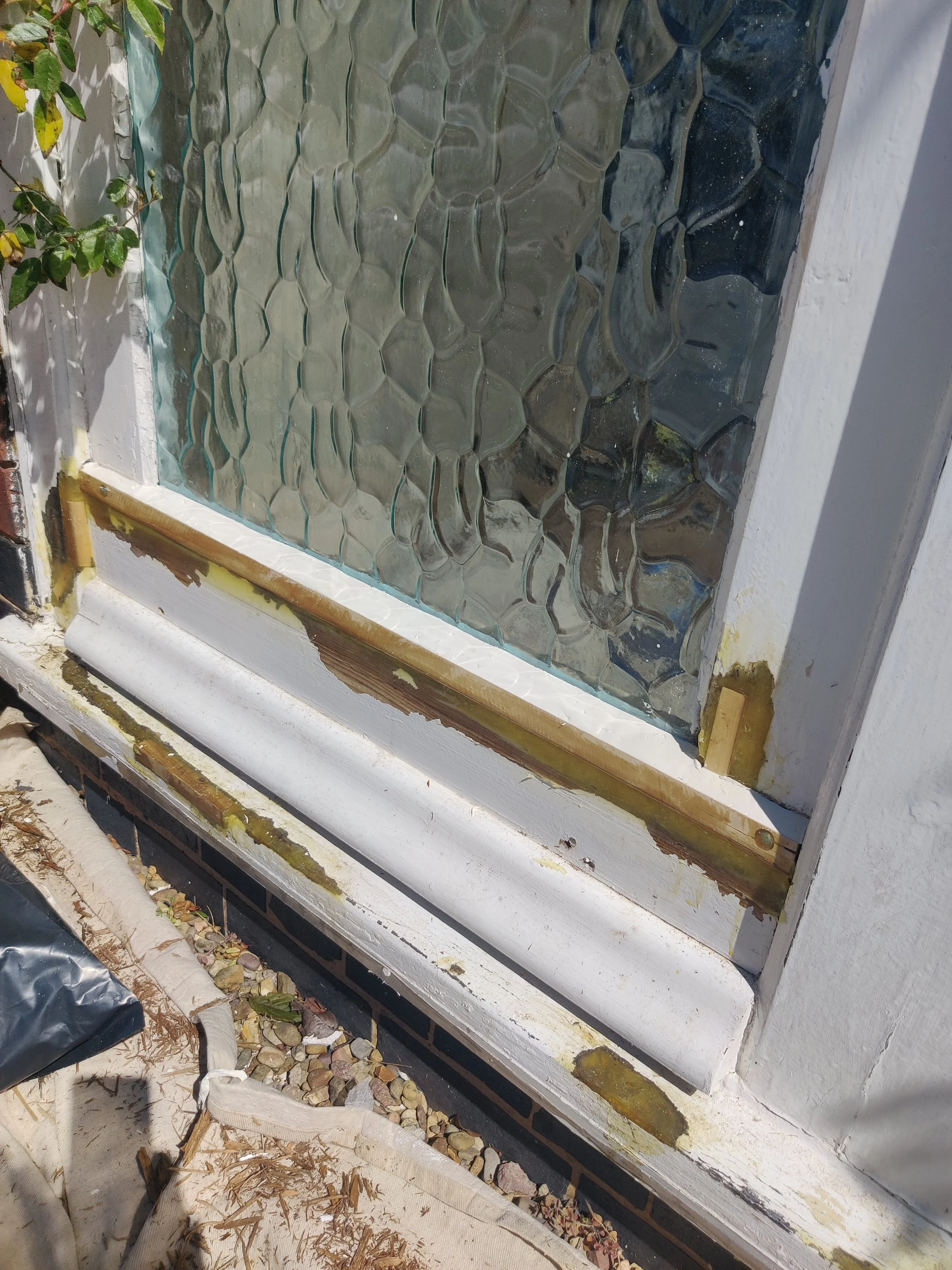 Close-up view of a window with textured glass panels and a white painted window sill. The window is being repaired or renovated, with visible tools, wood, and debris around. epoxy dryflex shaped to create original details