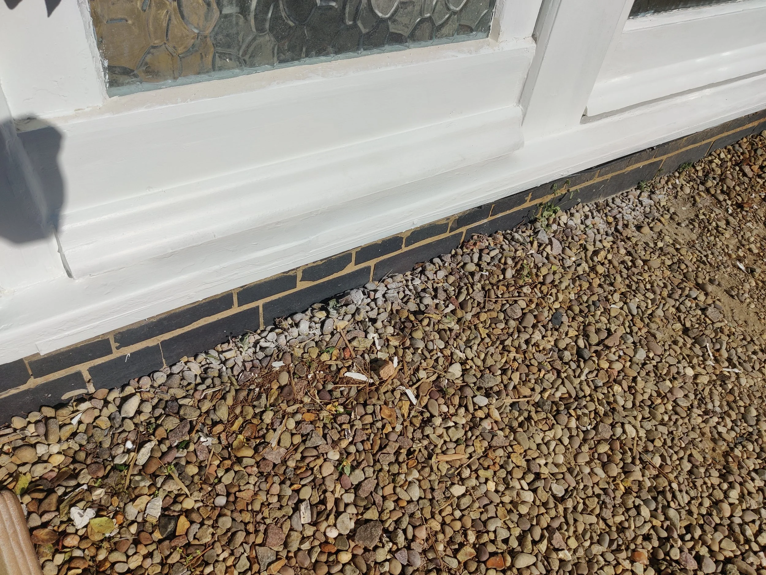 Close-up of a white framed window set in a brick wall, with small gravel stones on the ground below and tiny green plants growing among the stones.