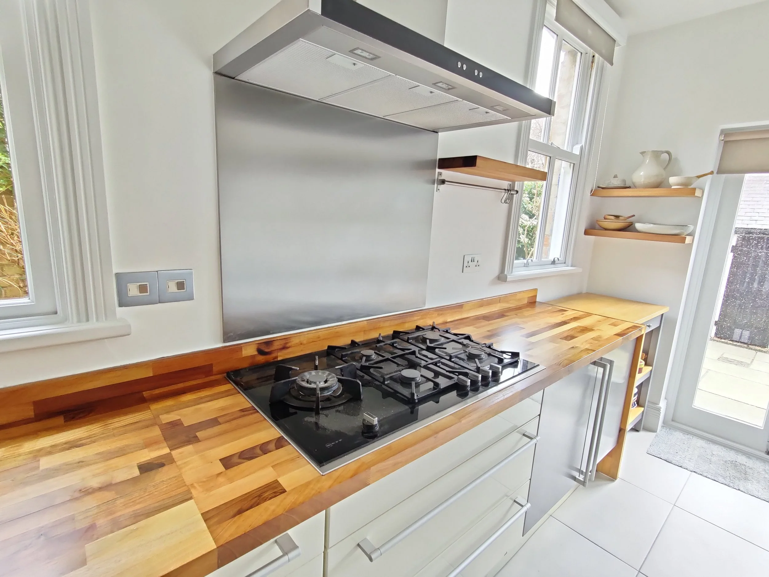 A kitchen with a freshly restore wooden countertop, gas stove, stainless steel range hood, white cabinets, and open shelves holding dishes, with windows and a door leading outside.