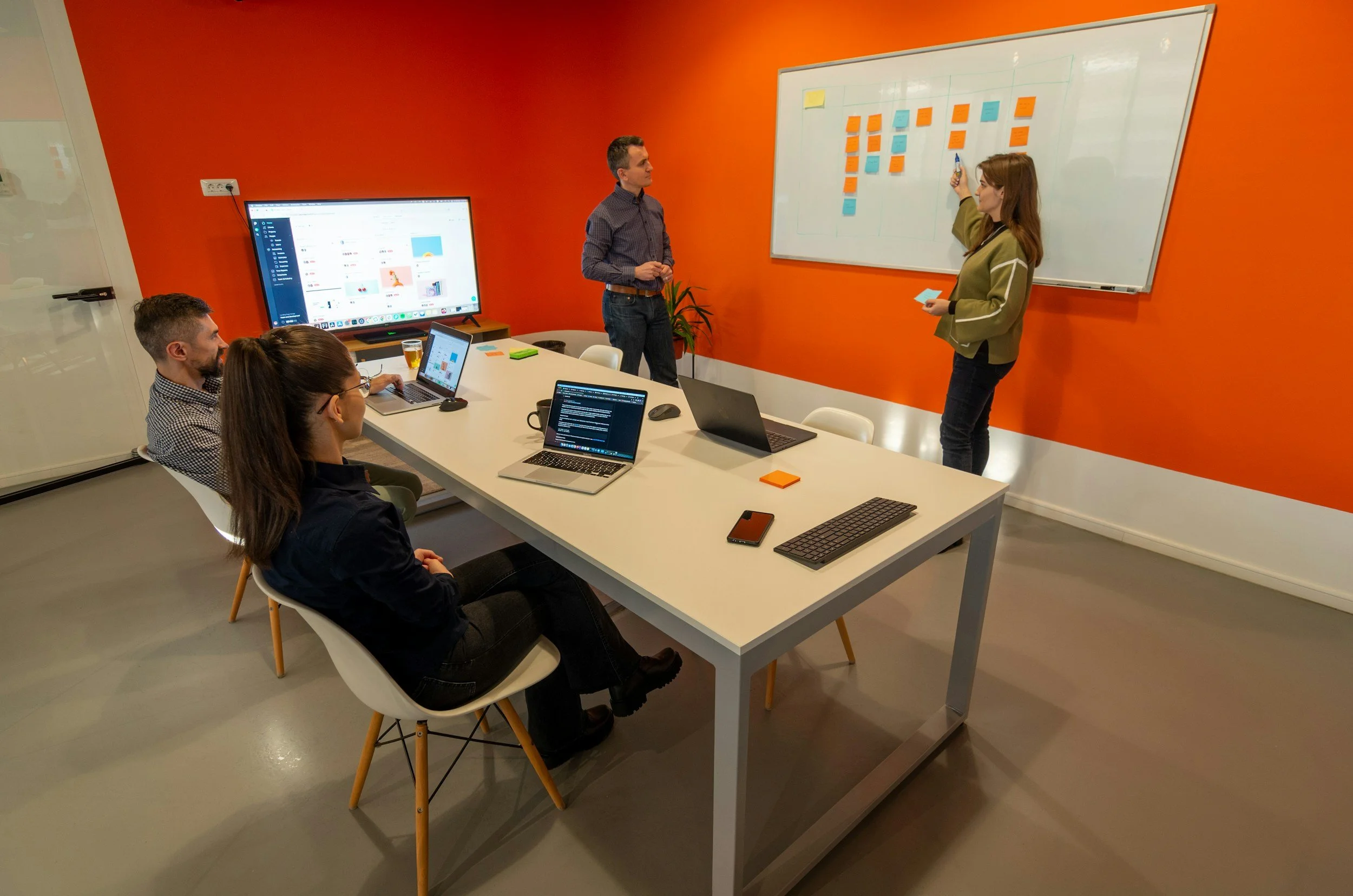 A business meeting in a conference room with five people, showing a woman presenting on a whiteboard with sticky notes, three laptops open on the table, and a large screen displaying a presentation, with a bright orange wall in the background.