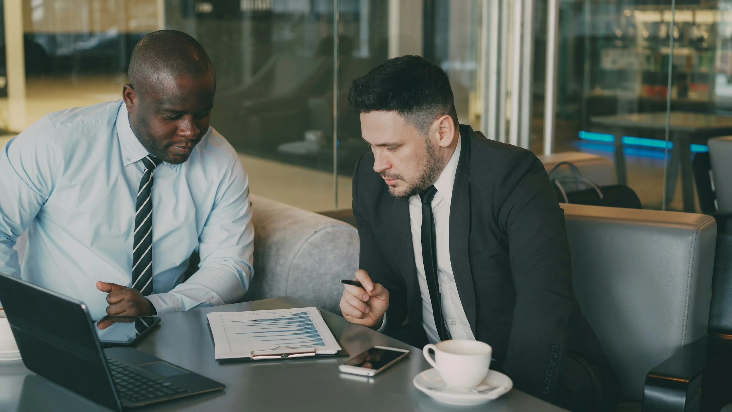 Two business professionals, one African American and one Caucasian, in formal attire, are seated at a table in a modern office or conference room. They are looking at a smartphone together, with documents and a laptop on the table.