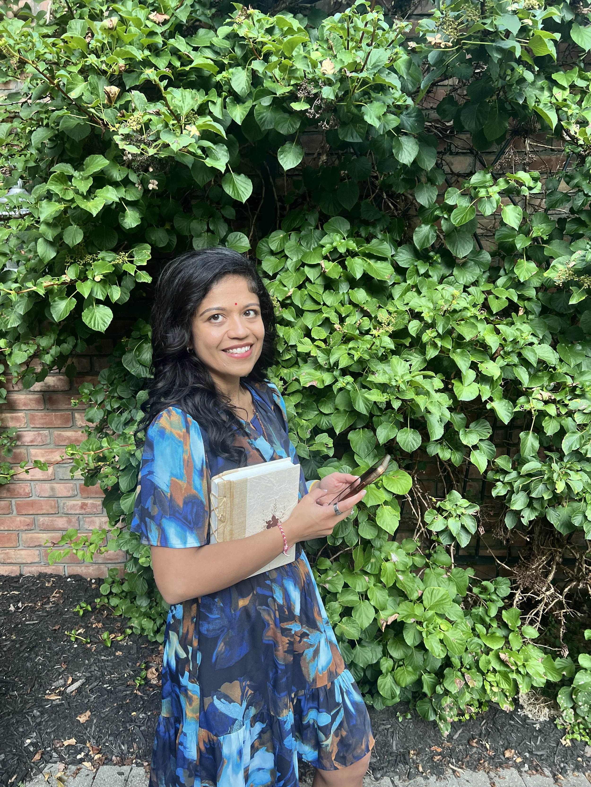 A woman (Nishi) with dark, wavy hair and a red bindi on her forehead stands outdoors in front of a lush, green leafy bush and a brick wall. She is smiling, holding a book and a smartphone, and wearing a blue dress.