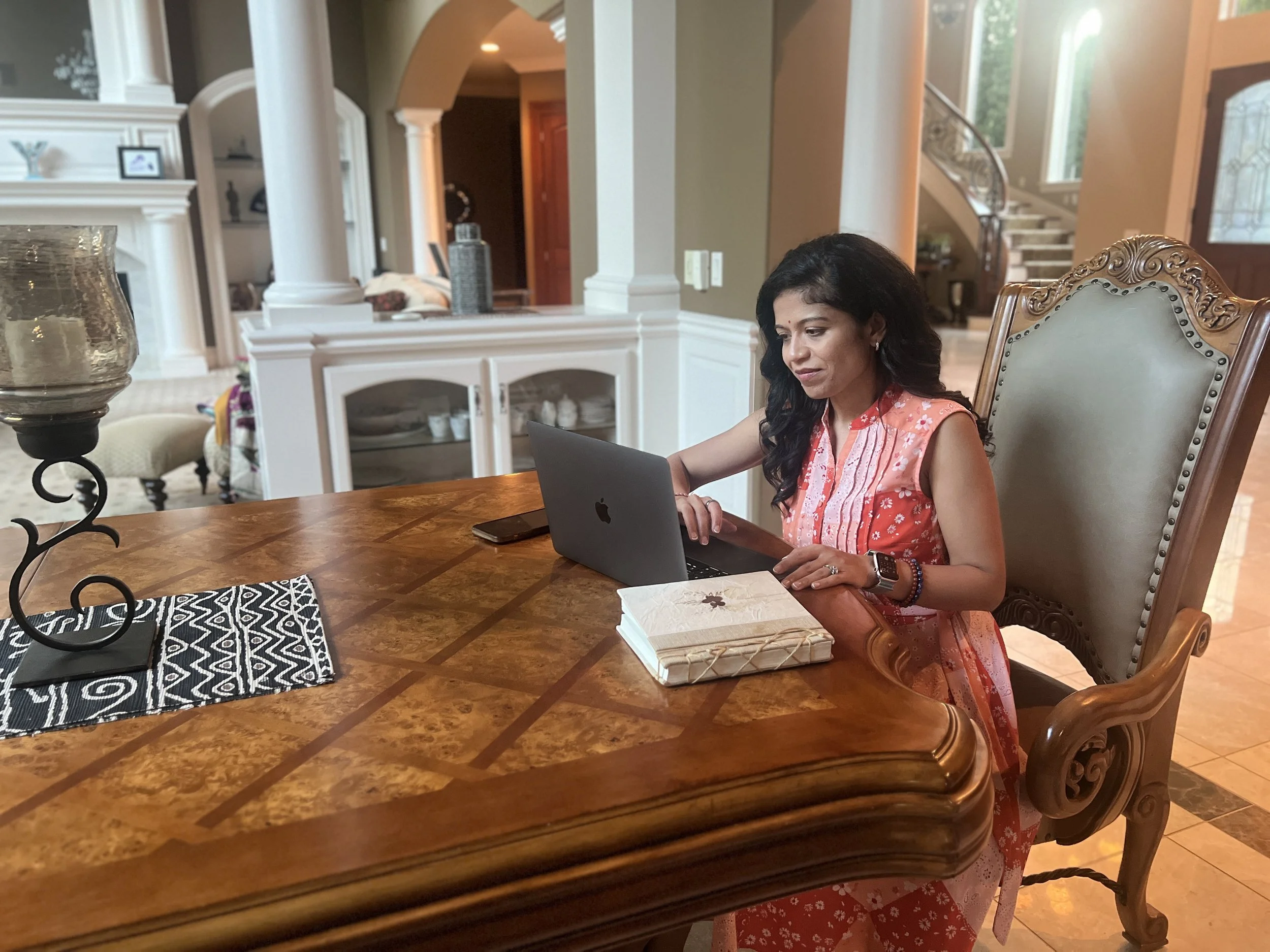 Nishi Jani Rajguru, sitting on a table inside appearing to work on her laptop. Has her phone and her notebook next to her.