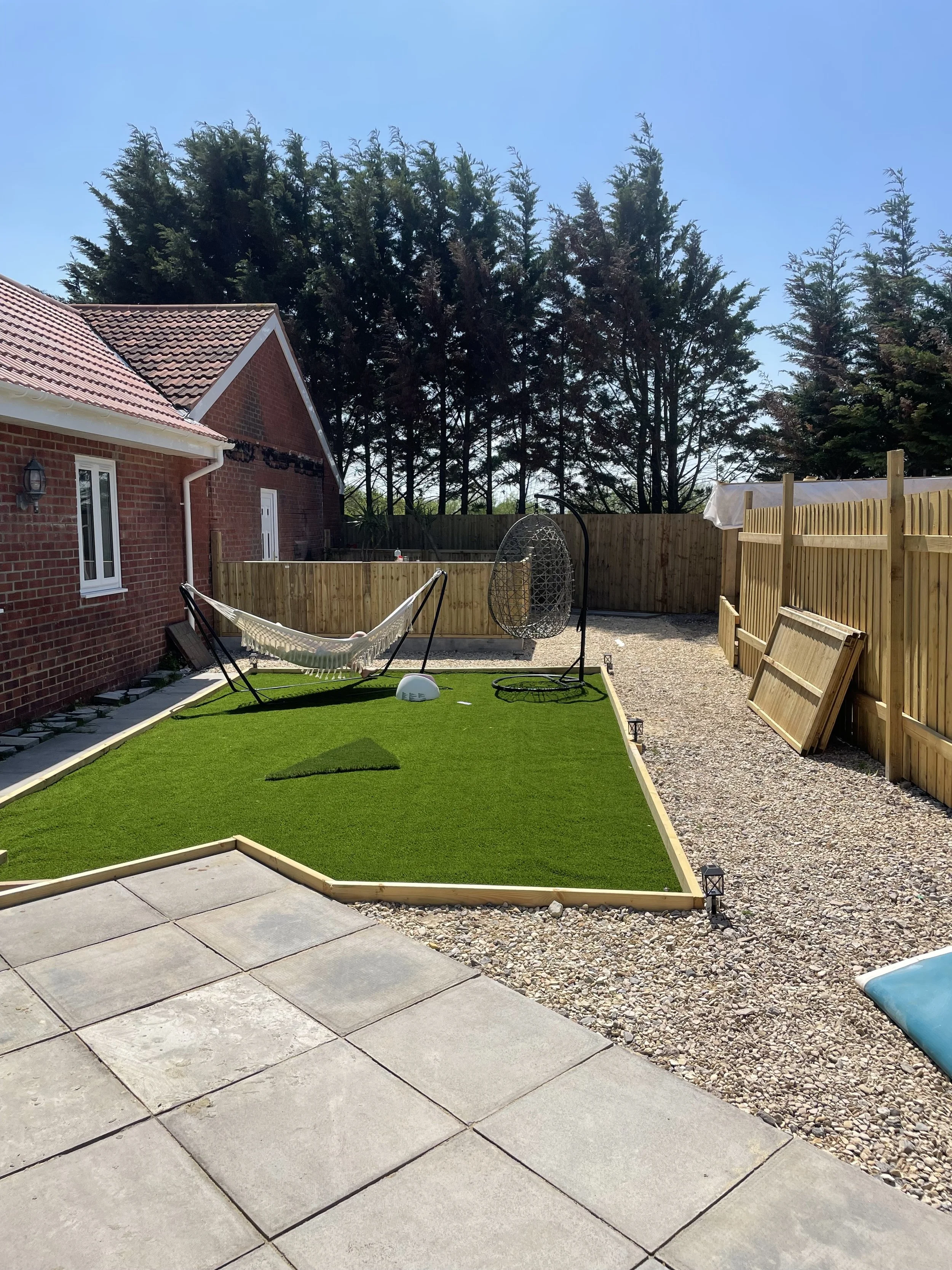 A backyard with freshly laid green artificial turf, a hammock suspended on a stand between a house and the yard, a hanging egg chair, a fenced area with a gravel ground, and a wooden fence with a white tarp or cover on top by Britton Landscaping in B