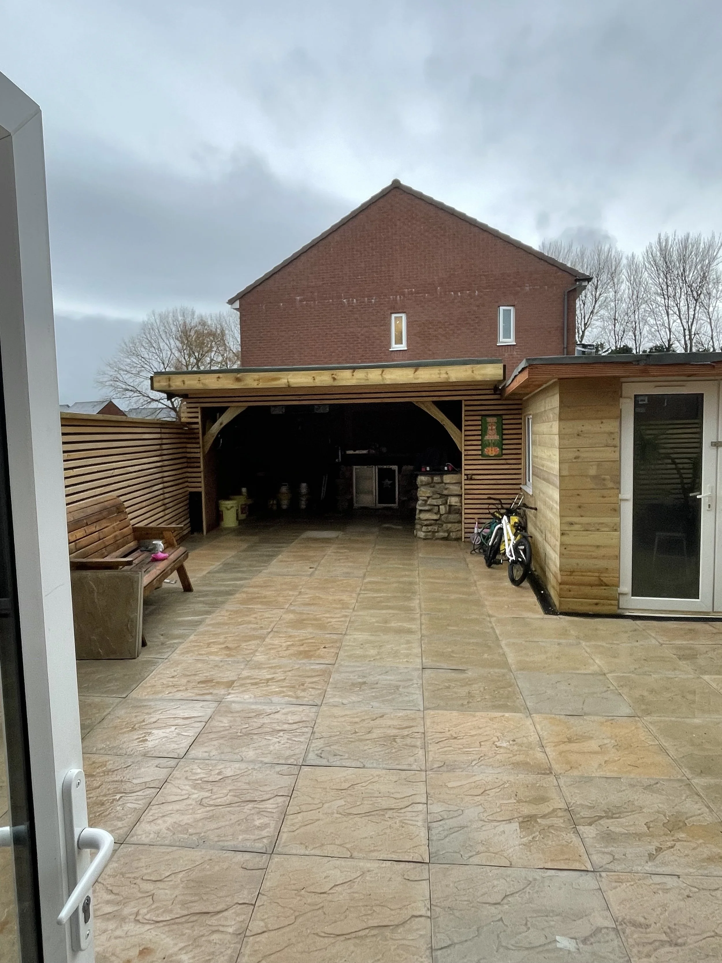 View of a backyard patio with beige stone tiles, a wooden bench, a wooden shed by Britton Landscaping in Bridgwater, Taunton, Weston Super Mare, Highbridge and Burnham