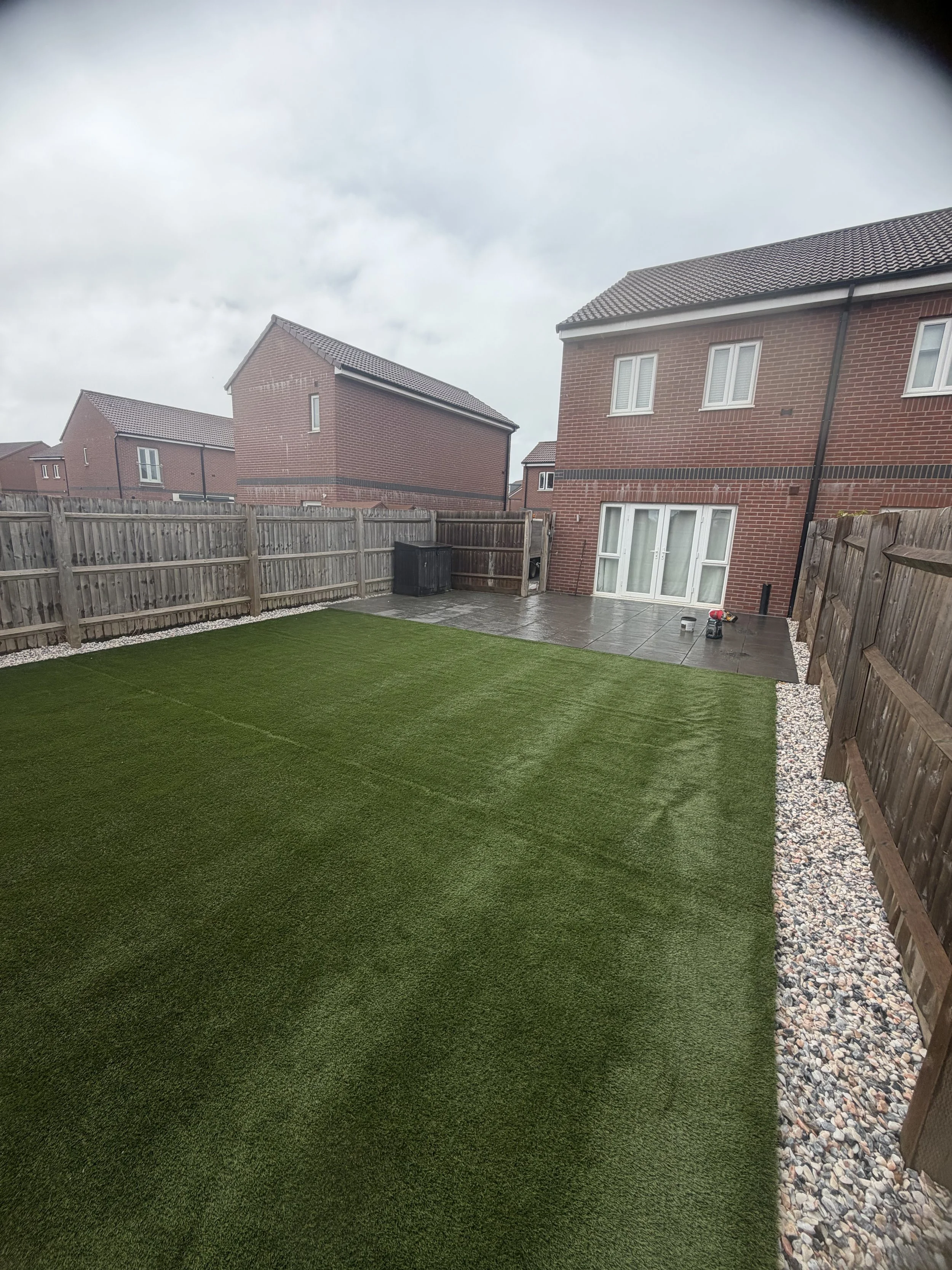 Backyard with artificial grass, wooden fence, and a brick house with sliding glass doors. Artificial turf installation by Brittons Landscaping in Taunton