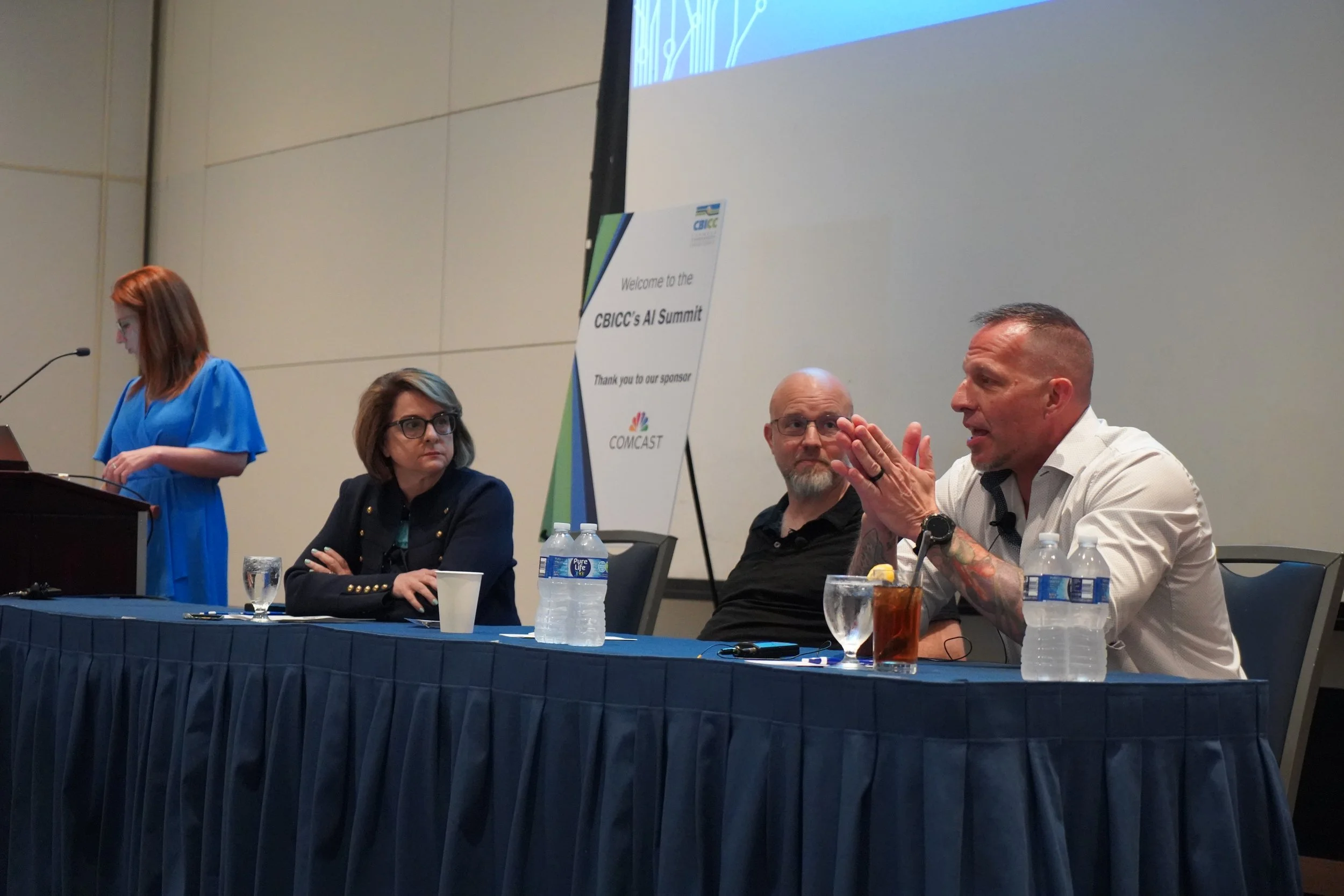 Four people sitting at a panel table during a conference with a woman standing at a podium speaking. The conference is the CBICC's AI Summit, sponsored by NBC and Comcast.