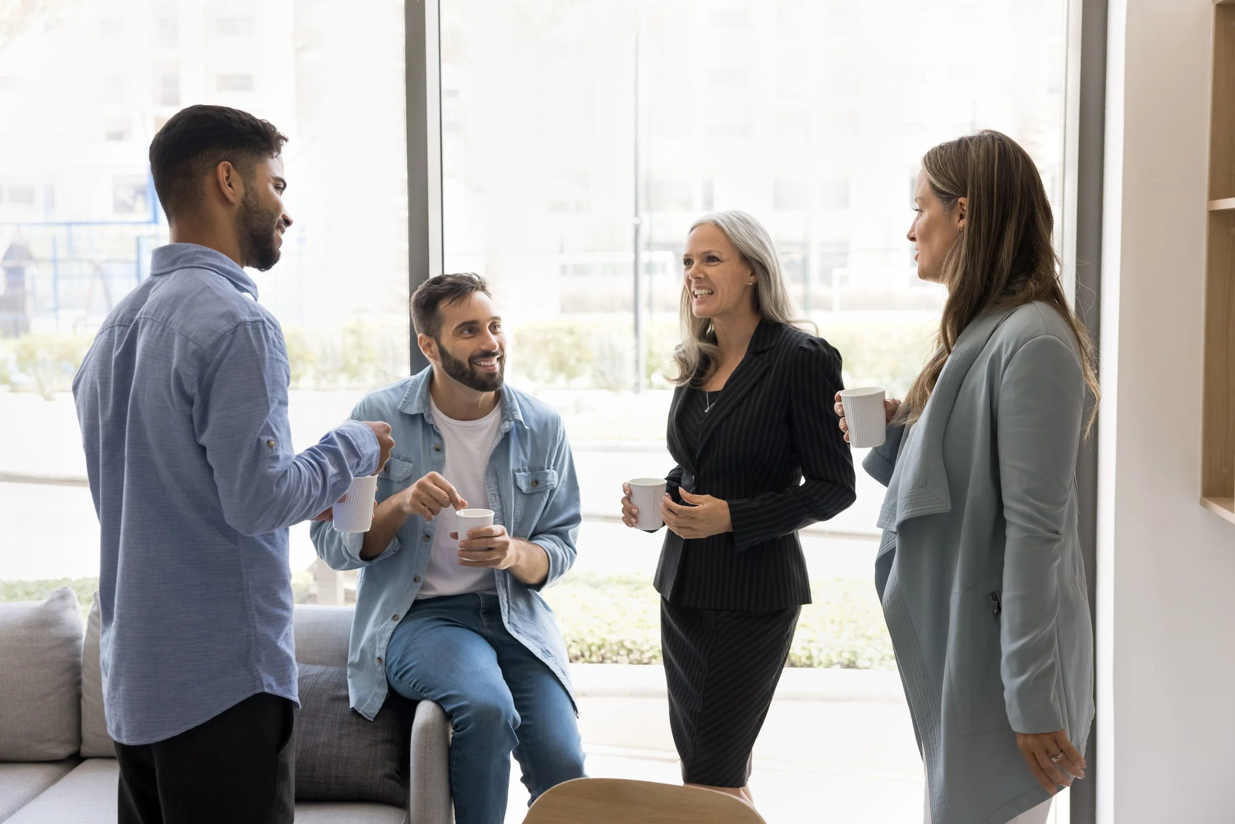 Four people having a conversation with coffee cups in a bright office space near large windows.