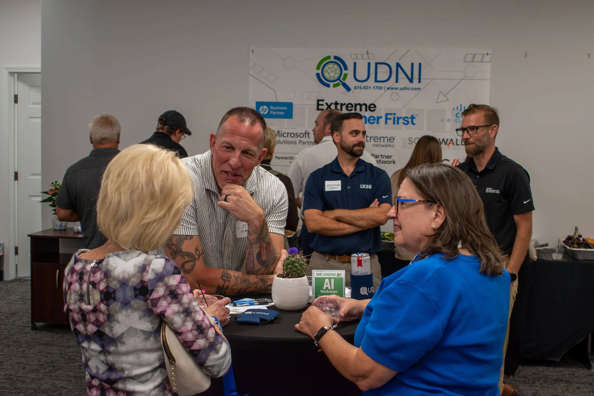 Bill Sell speaking with two women at a networking event in UDNI's Altoona, PA office, with a branded partner banner in the background.
