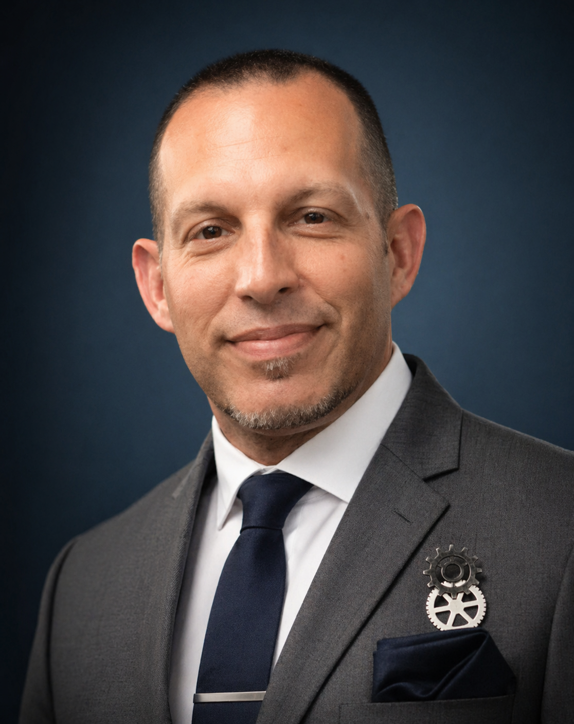 Bill Sell in a gray suit and white shirt, wearing a navy blue tie, with steampunk style gear lapel pin, smiling against a dark blue background.