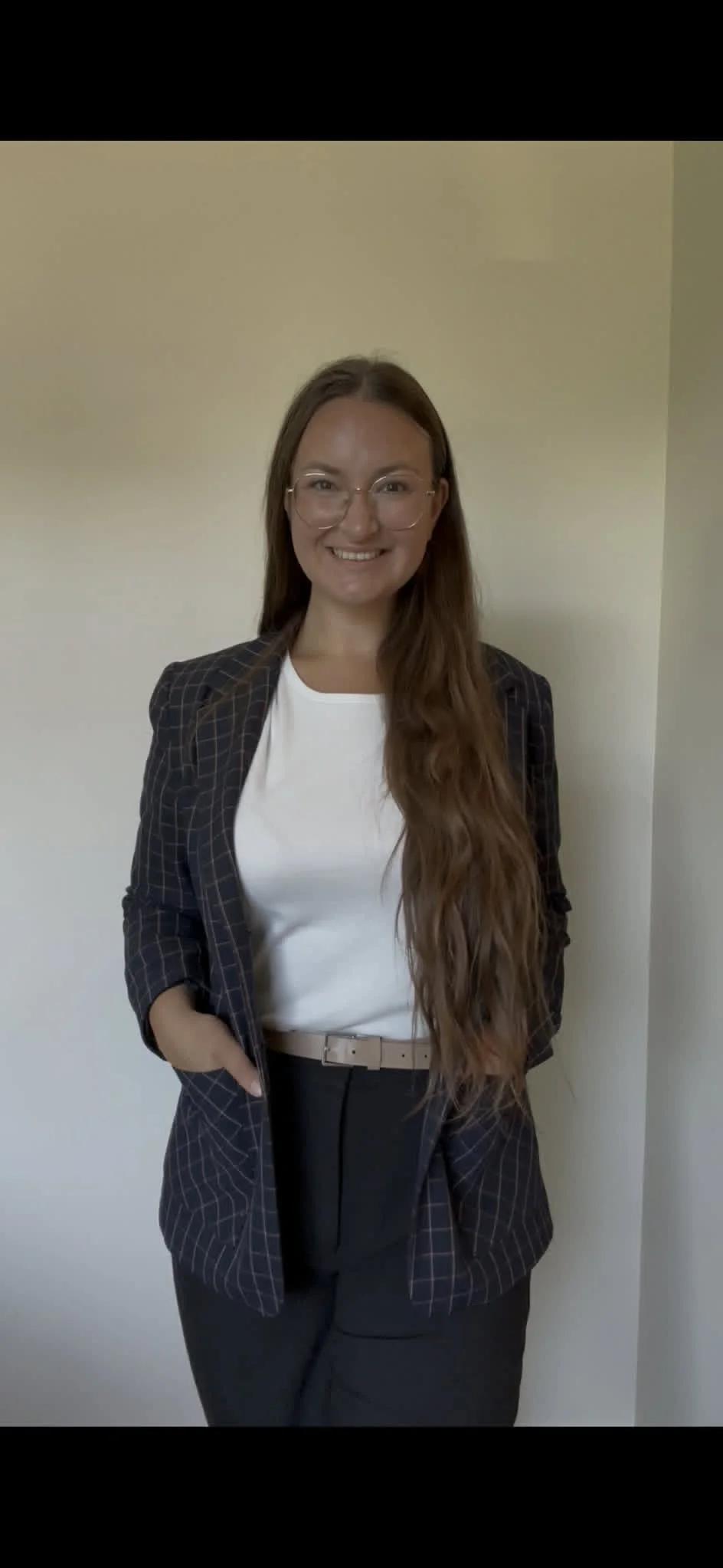 Young woman with long wavy brown hair, glasses, wearing a black plaid blazer over a white blouse, smiling, standing against a plain wall.