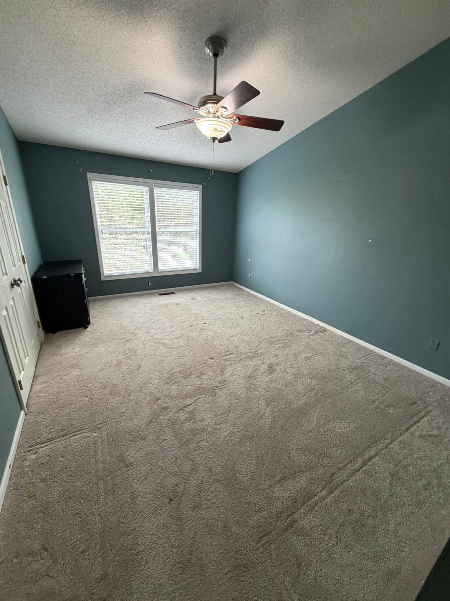 Empty room with beige carpet, blue walls, a window with blinds, a ceiling fan with lights, and a small black cabinet.