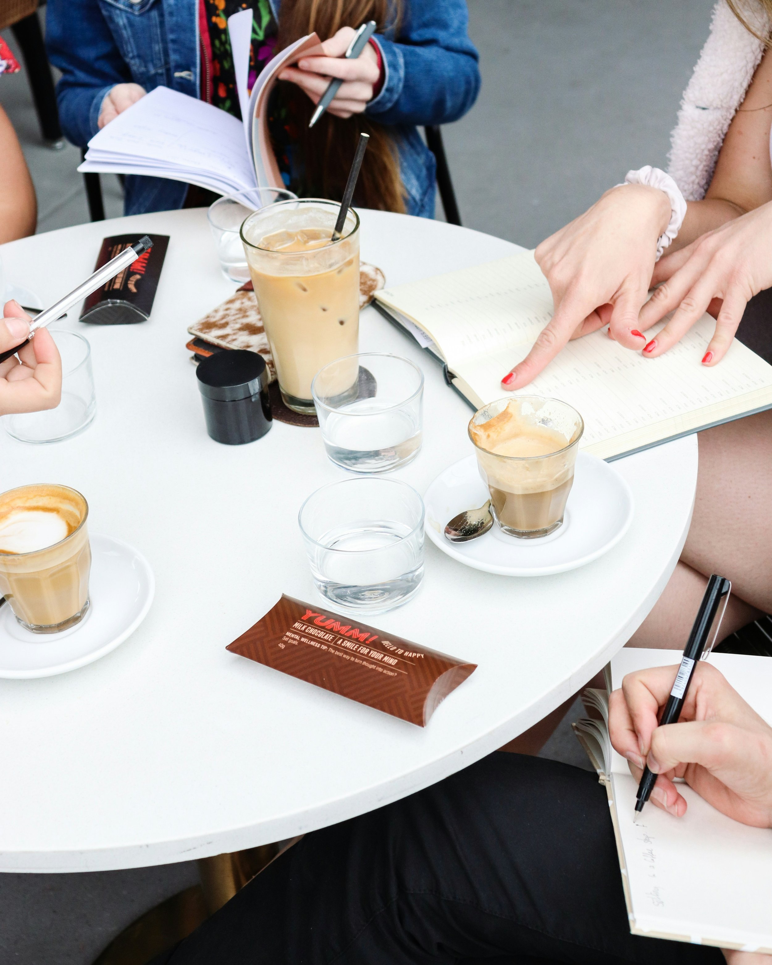A group of people sitting around a white table, with notebooks, pens, glasses of water, and iced coffee drinks. One person is pointing at their notebook with a red manicure. Someone else is holding a pen and writing.
