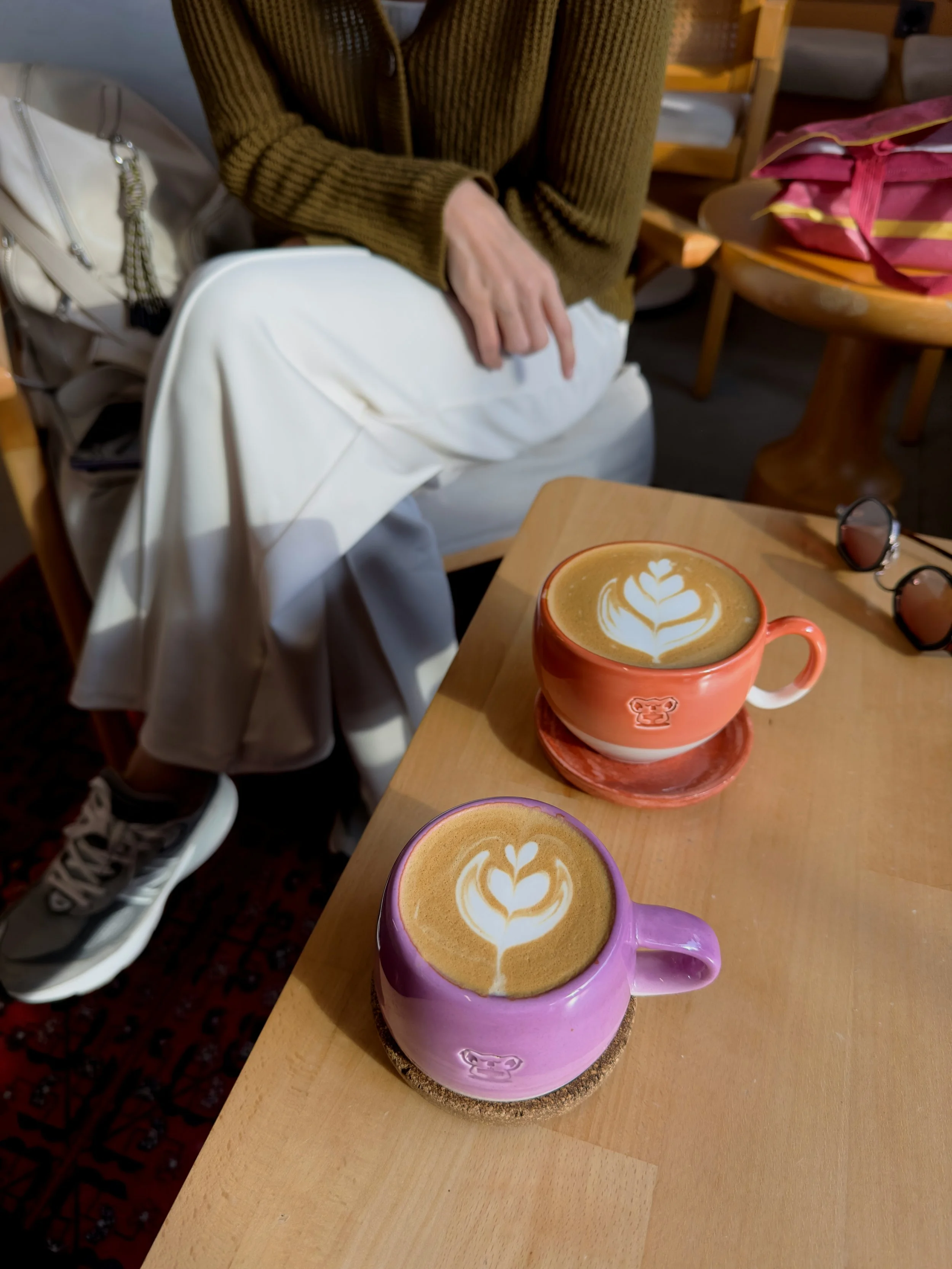 Two colorful mugs of latte with latte art on a wooden table, with a person sitting nearby, a pair of sunglasses on the table, and a pink bag in the background.