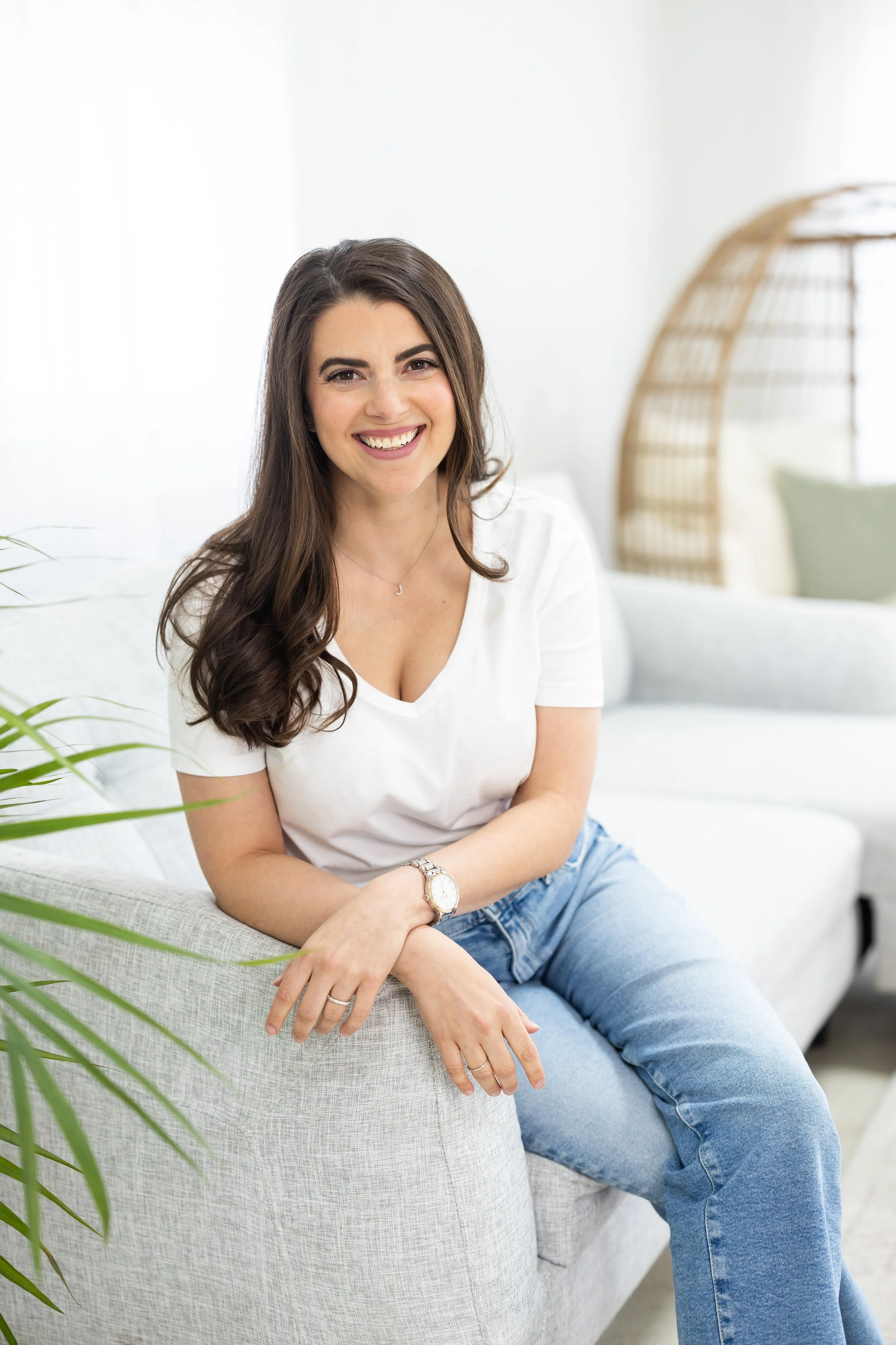 A young woman with long brown hair, wearing a white t-shirt and blue jeans, smiling and sitting on a beige sofa in a bright, minimalist room with a wicker lamp in the background and some green plants.