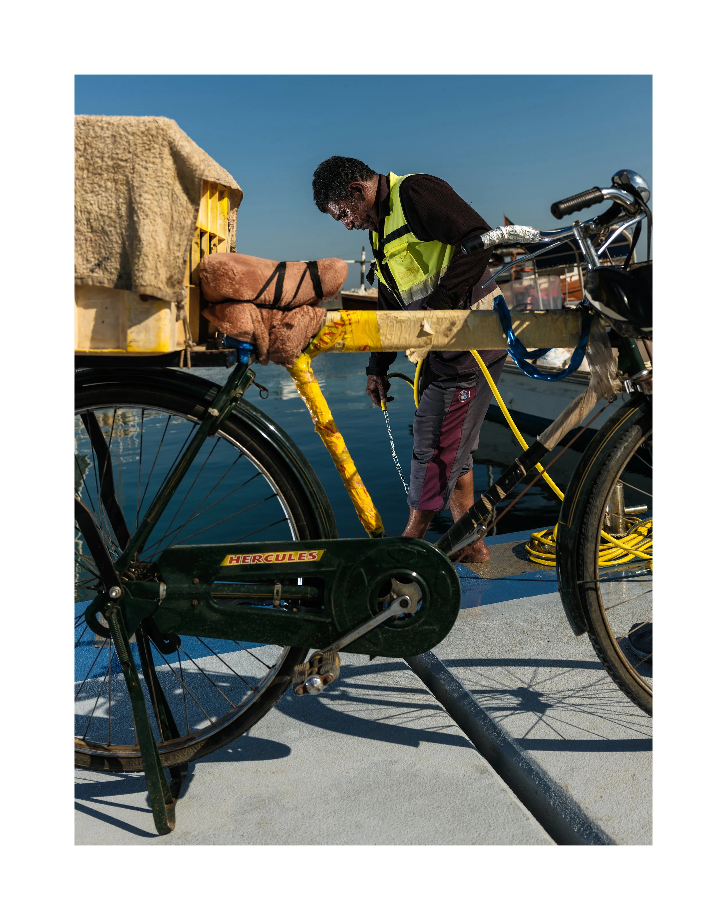 A man wearing a yellow safety vest cleaning a bicycle on a dock by the water, with boats and a clear blue sky in the background.