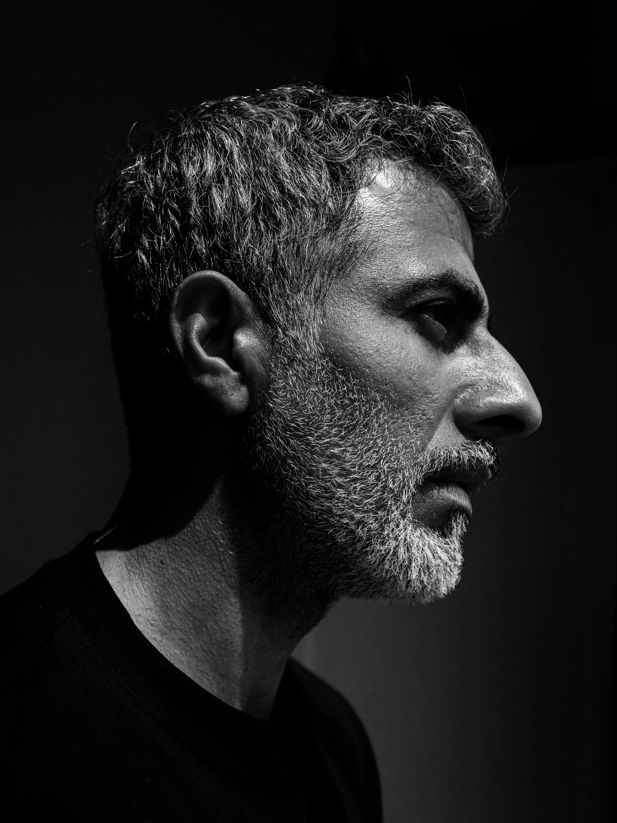 A black and white profile portrait of an older man with gray curly hair, a beard and mustache, wearing a dark shirt, against a dark background.
