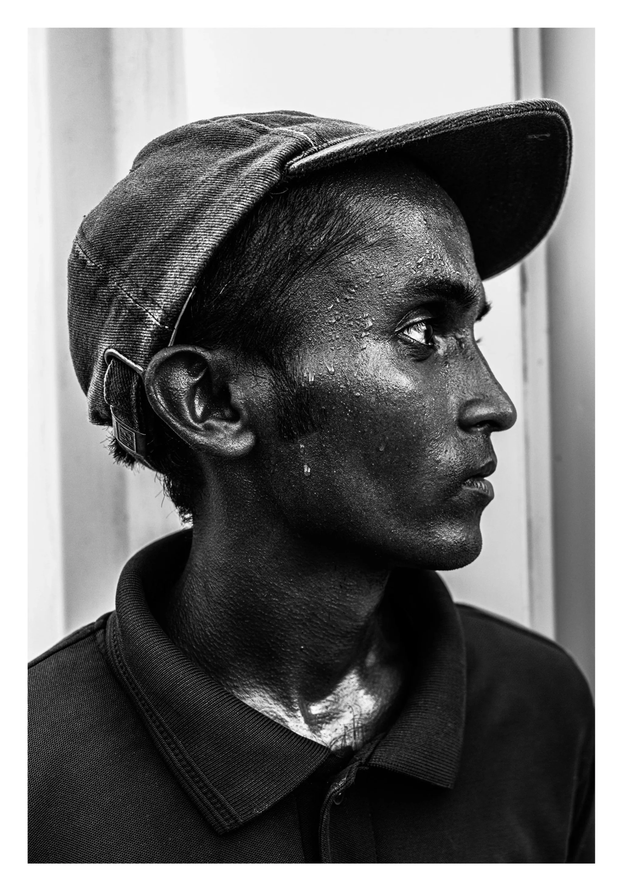 A black and white side profile of a man wearing a cap and a collared shirt, with sweat on his face.