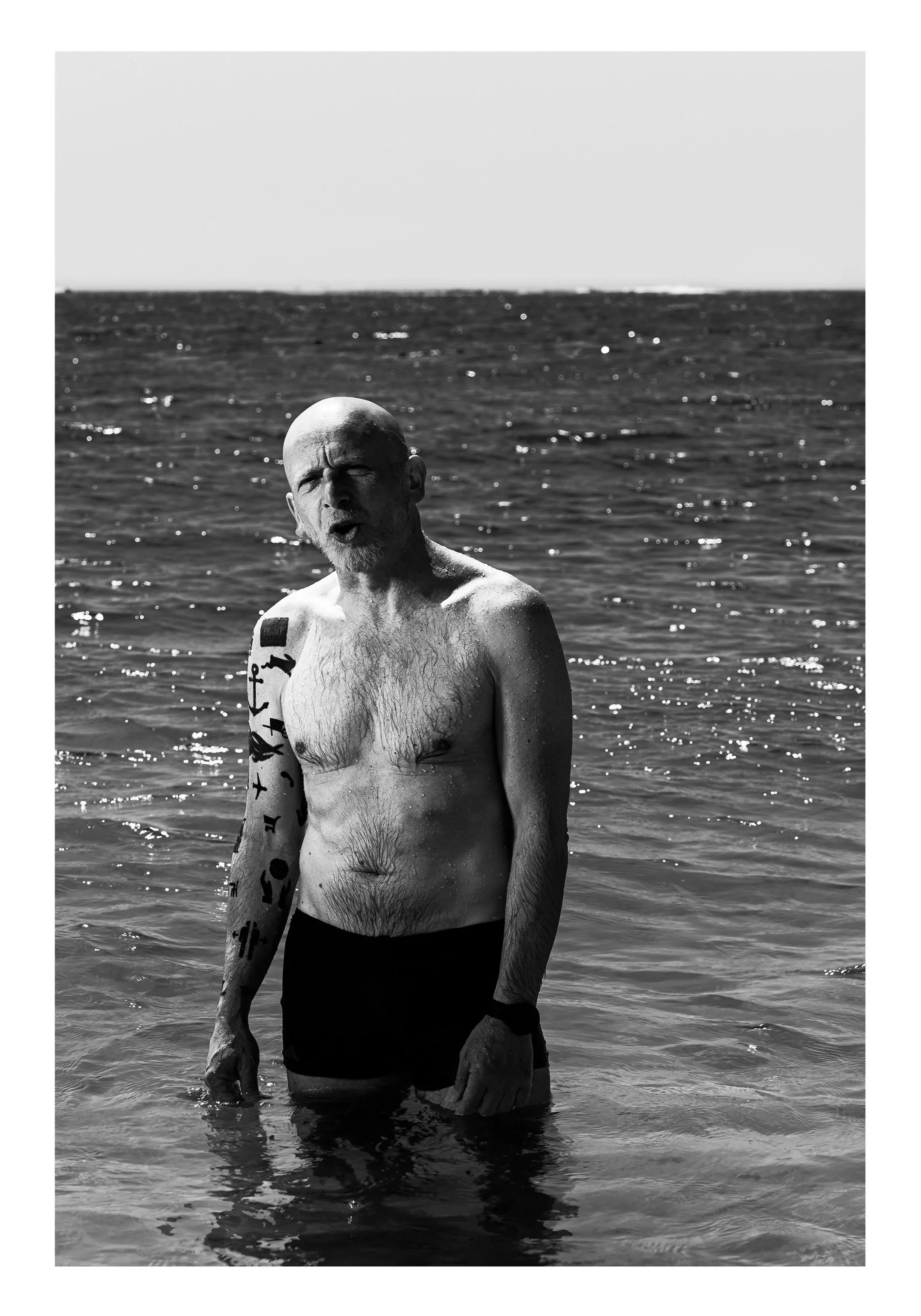 A black and white photo of a bald, bearded man with tattoos on his arms, standing in the ocean wearing dark shorts, with water reaching just above his knees. He appears to be looking at the camera with his mouth slightly open.