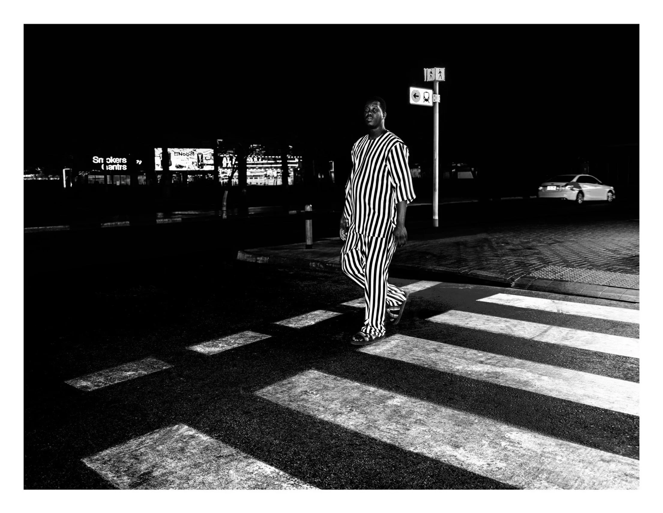 A man dressed in black and white striped clothing crossing a zebra crosswalk at night, with cars and illuminated signs in the background.