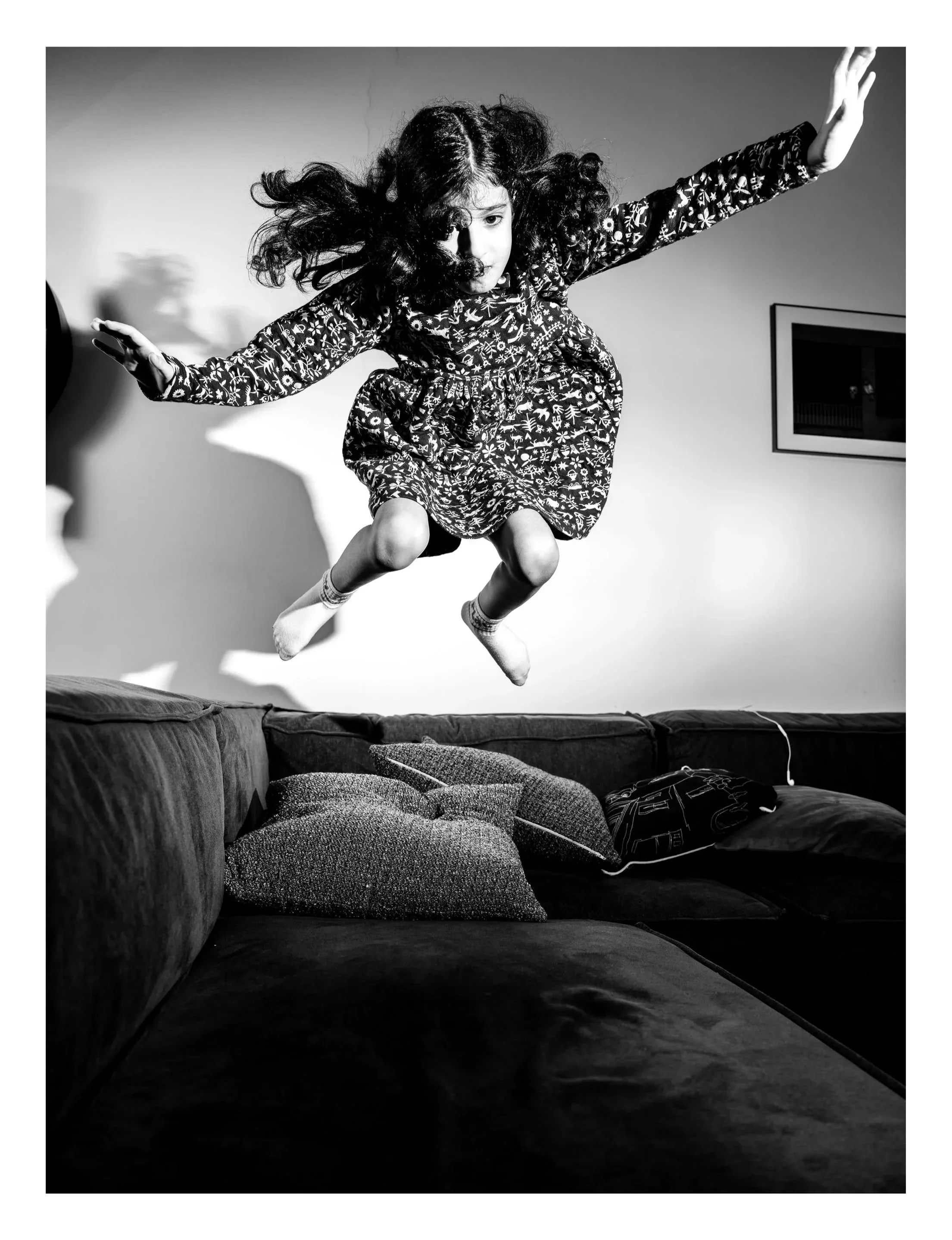 A young girl with curly hair is jumping onto a sofa inside a room. The room has a framed picture on the wall. The photo is in black and white.