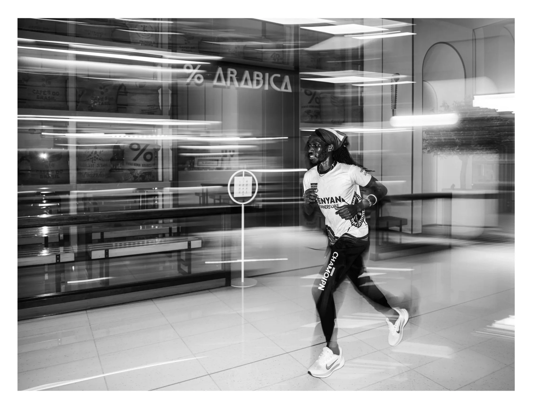 A man running indoors, wearing a white shirt with a Kenyan flag and the word Kenya, black pants, and white sneakers, with motion blur in the background.
