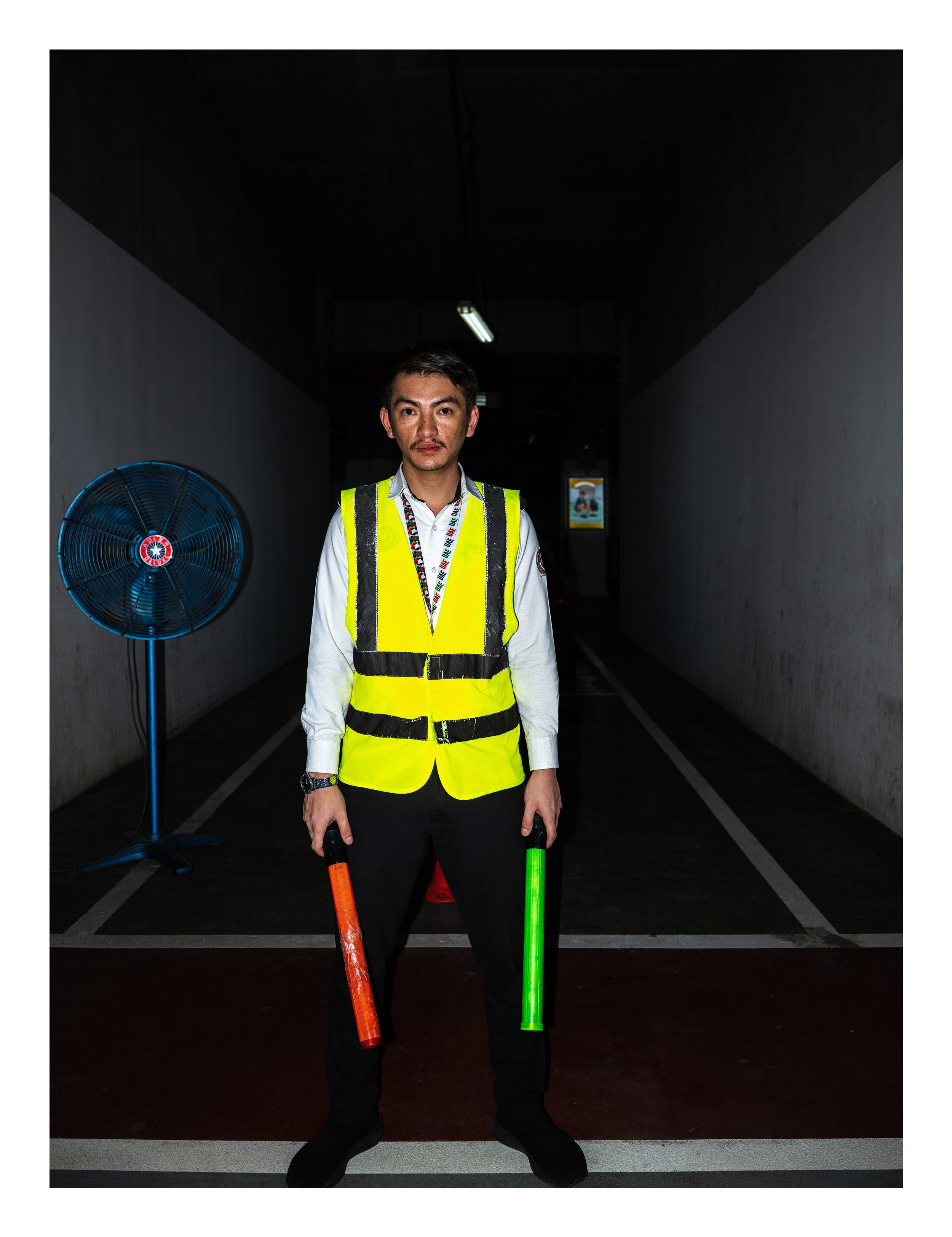 A man wearing a white shirt, yellow safety vest, and black pants standing in a dark corridor holding orange and green glow sticks. There is a blue fan on his left side and a screen in the background.