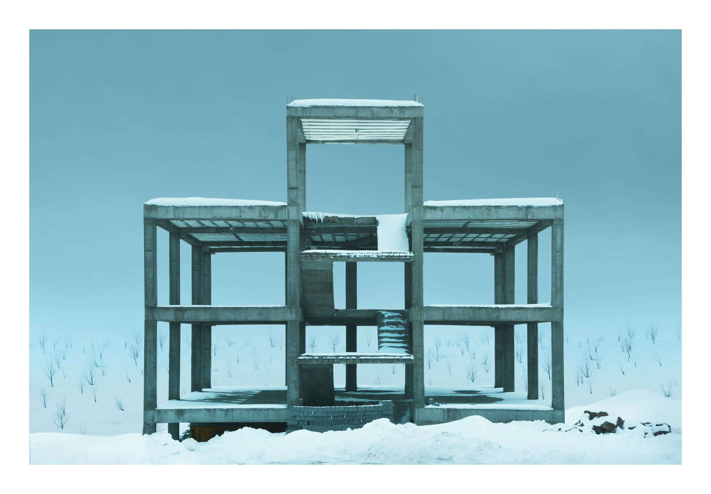 A partially constructed concrete building structure covered in snow, with a staircase in the middle and a snowy hill with small leafless trees in the background.