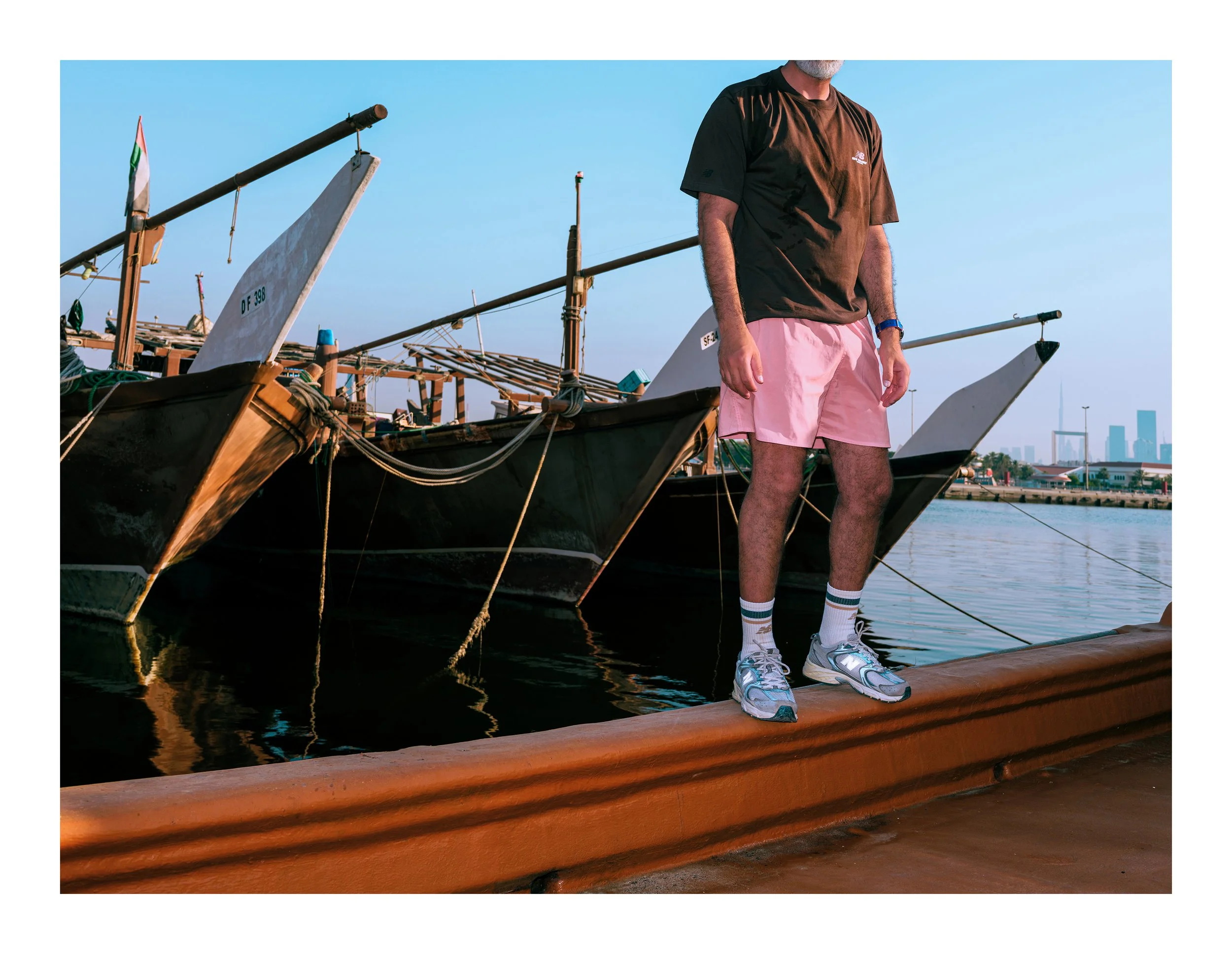 A man in black t-shirt, pink shorts, and sneakers standing on a wooden dock by boats docked at the harbor, with city skyline in the background.
